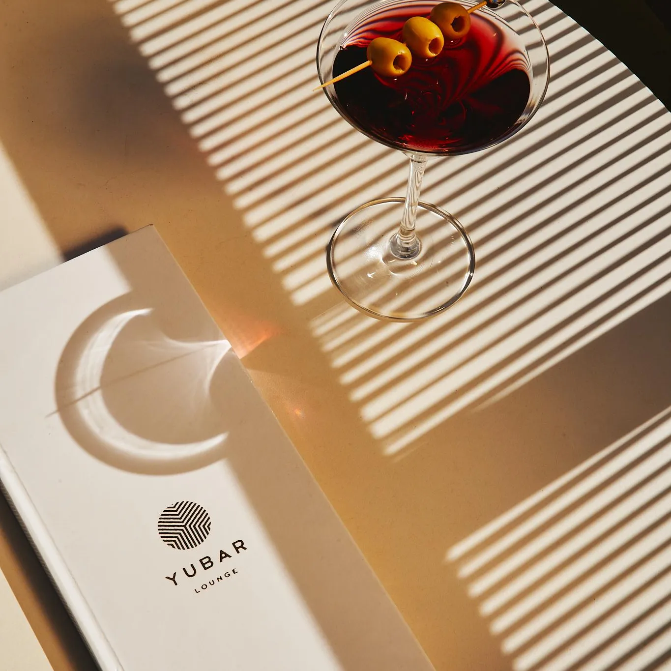 Un verre de cocktail avec des olives et une paille, posé sur une table en bois avec un menu "YUBAR LOUNGE" à côté