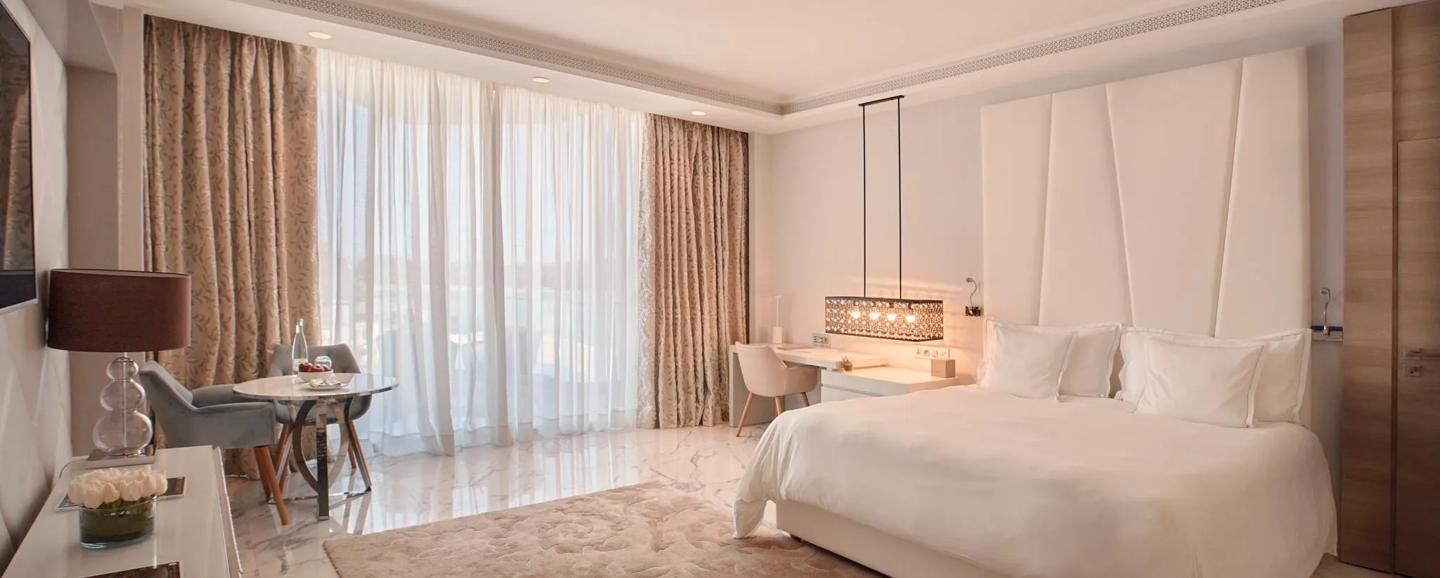 Habitación de hotel con una cama grande blanca, una mesita de noche con una lámpara, un rincón de salón cerca de una gran ventana con las cortinas abiertas