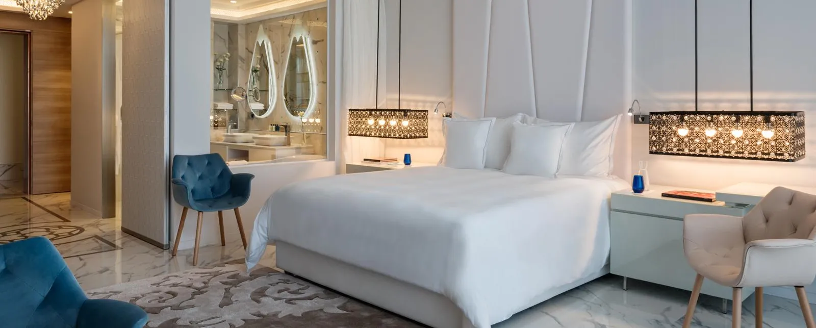 Habitación lujosa con cama blanca, dos sillones, mesita de noche y vista a un baño con espejos ovalados