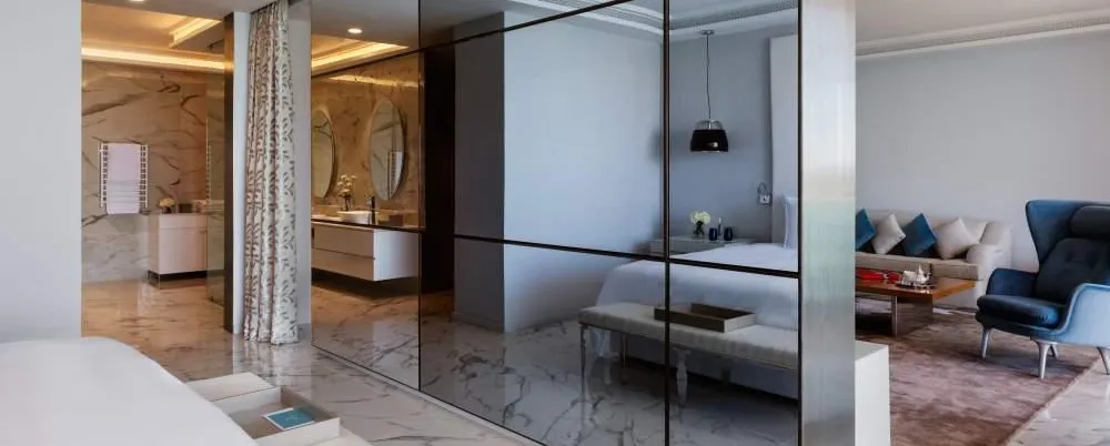 Interior moderno con un espejo que separa un dormitorio y una sala de estar, mostrando muebles blancos y un rincón de estar con sofá y sillón.
