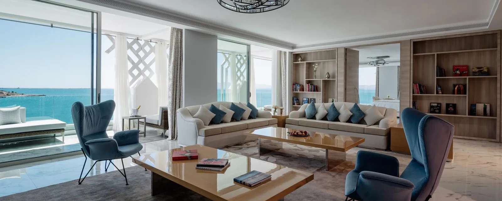 Salón moderno con vista al mar, que incluye un sofá, sillones, una mesa baja y estanterías llenas de libros