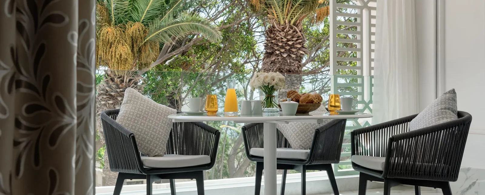 Mesa redonda con cuatro sillas de ratán negro y cojines grises, vista desde una puerta abierta, con plantas y elementos decorativos sobre la mesa en el fondo