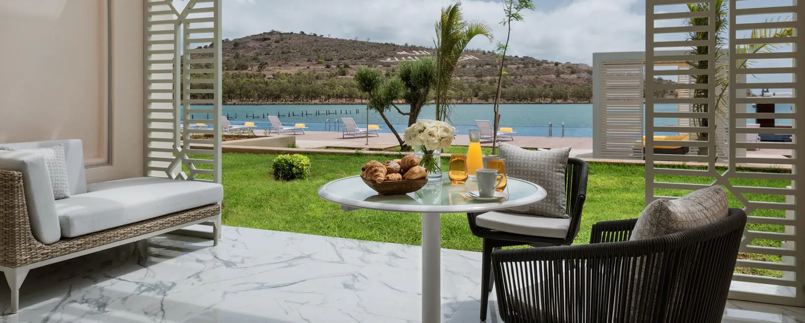 Terraza con mesa, sillas y sofá que da a un césped y un lago con tumbonas y montañas de fondo