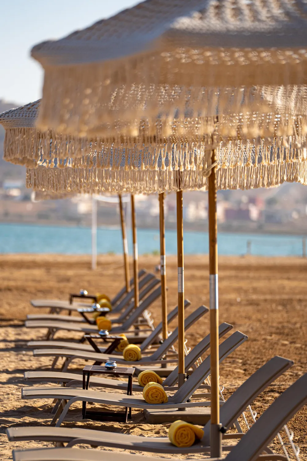 A row of deck chairs with yellow towels under straw umbrellas on a beach by the water