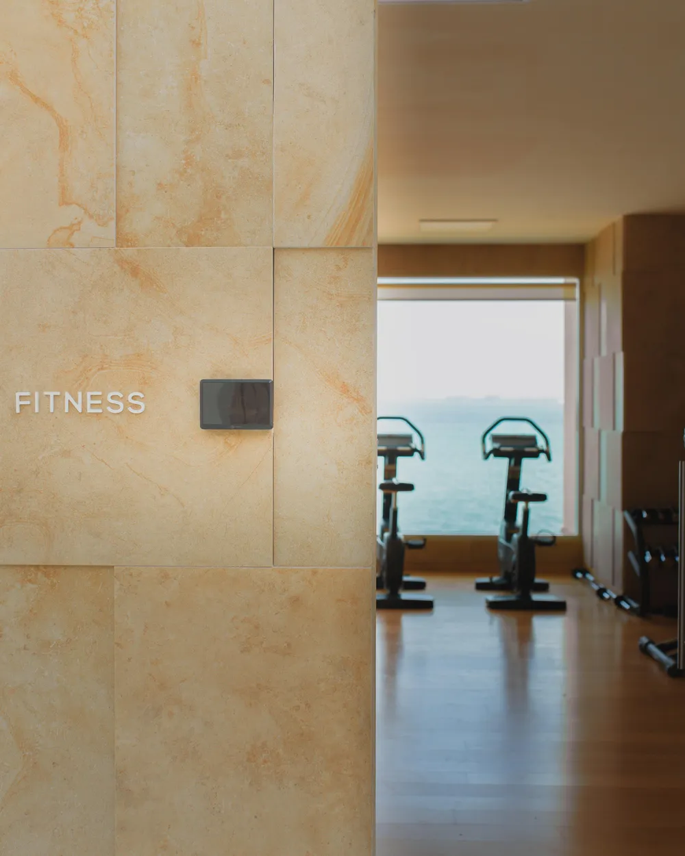 Vista de un espacio de fitness con bicicletas de ejercicio, visible a través de una puerta abierta, y una pared de piedra con la inscripción "FITNESS"