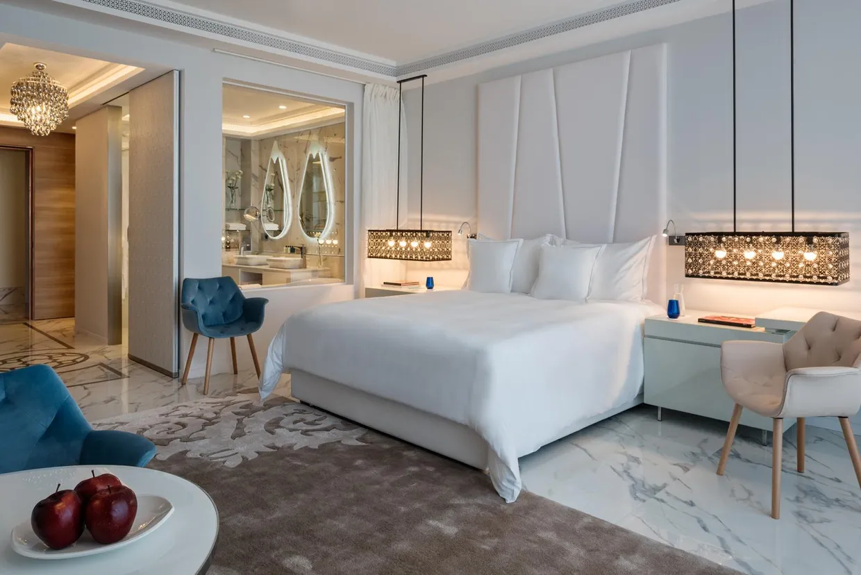 Luxurious room with white bed, two armchairs, bedside table, and view of a bathroom with oval mirrors.
