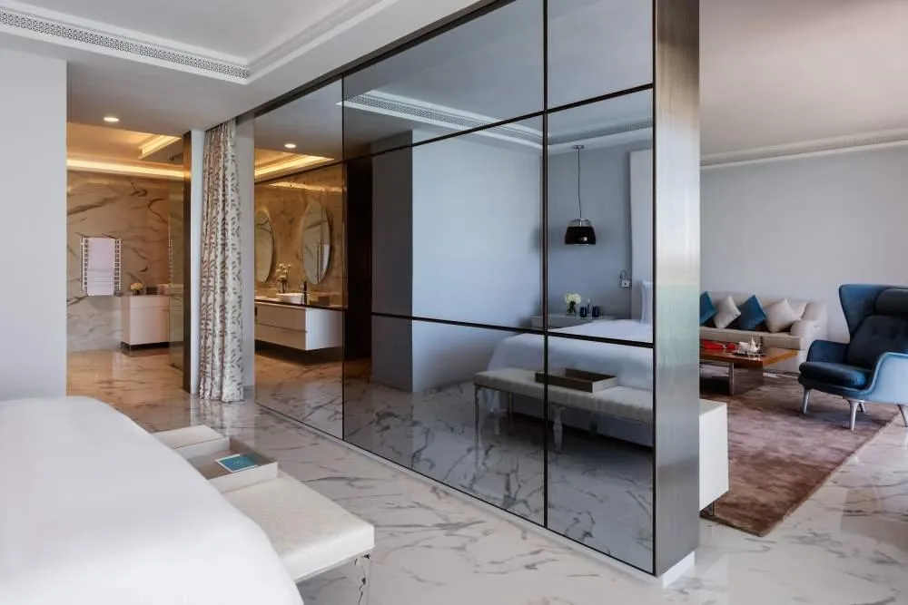 Modern interior with a mirror separating a bedroom and a living room, featuring white furniture and a seating area with a sofa and armchair.