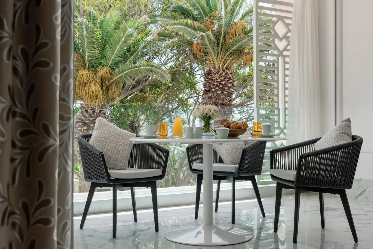 Round table with four black rattan chairs and gray cushions, seen from an open door, with plants and decorative elements on the table in the background.