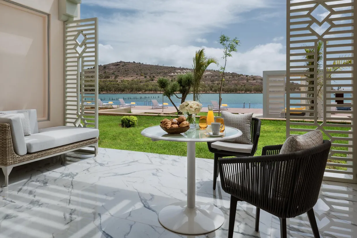 Terrace with table, chairs, and sofa overlooking a lawn and a lake with sun loungers and mountains in the background.