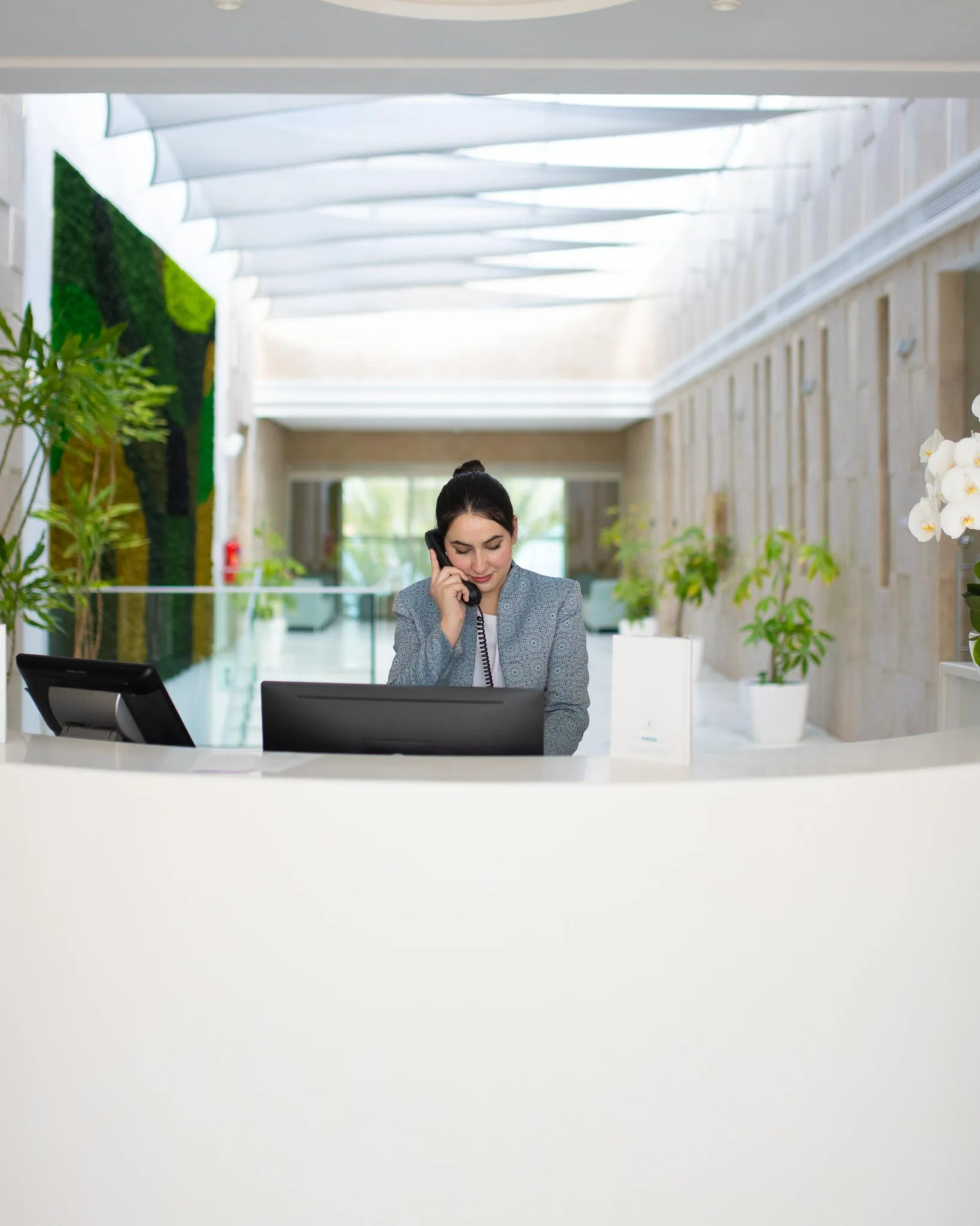 A woman in a suit talks on the phone behind a reception desk with plants and a computer.