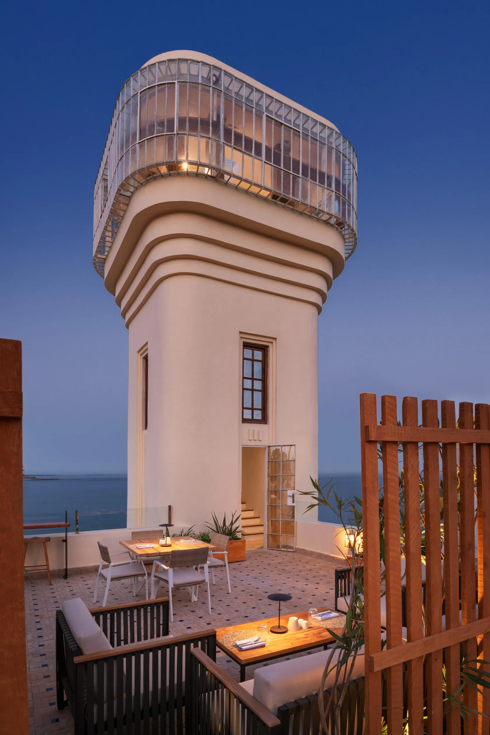 Un bâtiment blanc avec une terrasse vitrée en forme de dôme surplombe la mer, avec des tables et des chaises disposées sur une terrasse en pierre