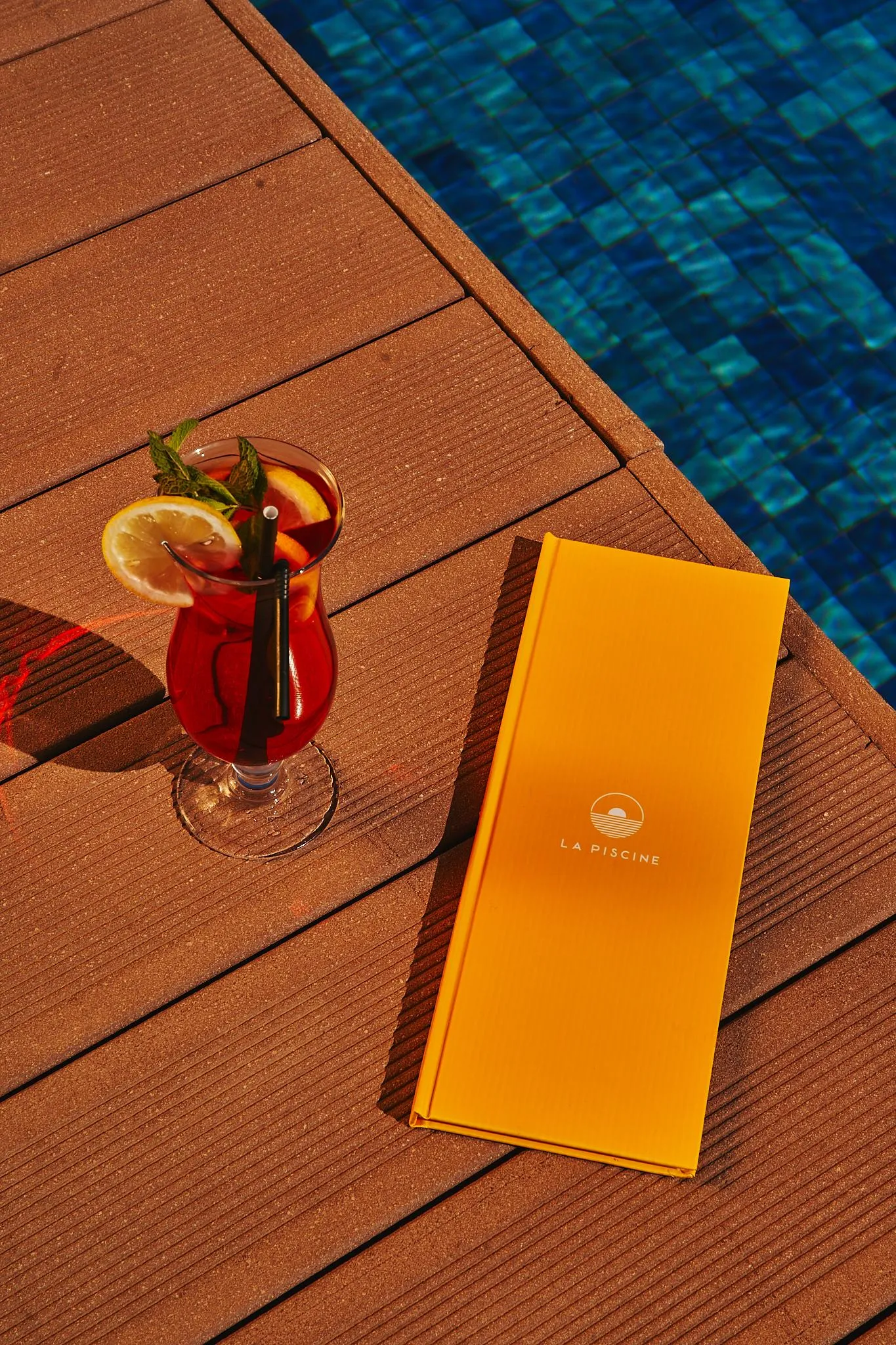 A glass of red drink with an orange slice and a straw, an orange menu, and a blue pool in the background.