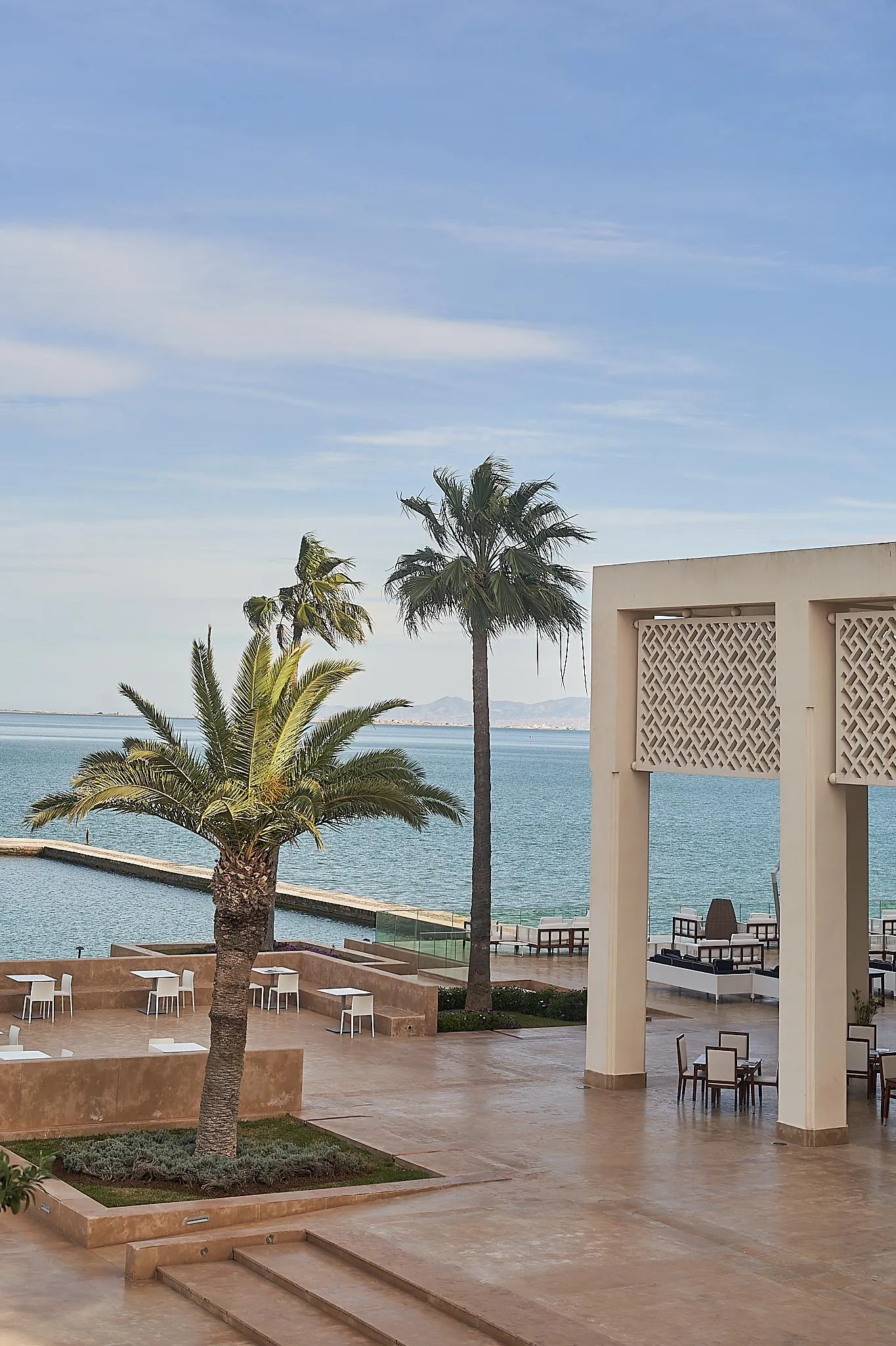 Terrasse avec tables et chaises près de la mer, bordée de palmiers et d'une structure architecturale avec des motifs géométriques
