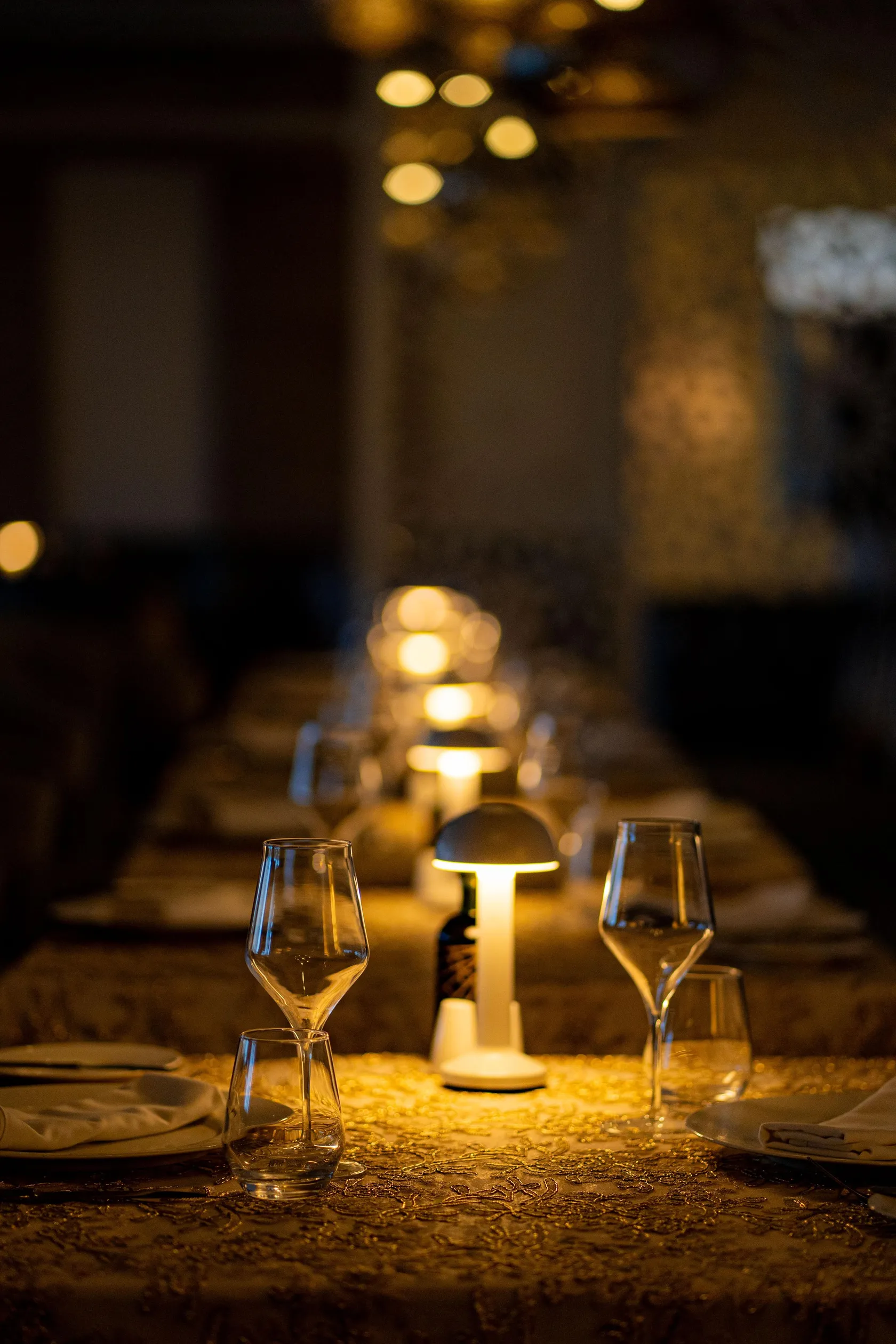 Table set with wine glasses, tablecloth, and candles, lit by lamps in the background