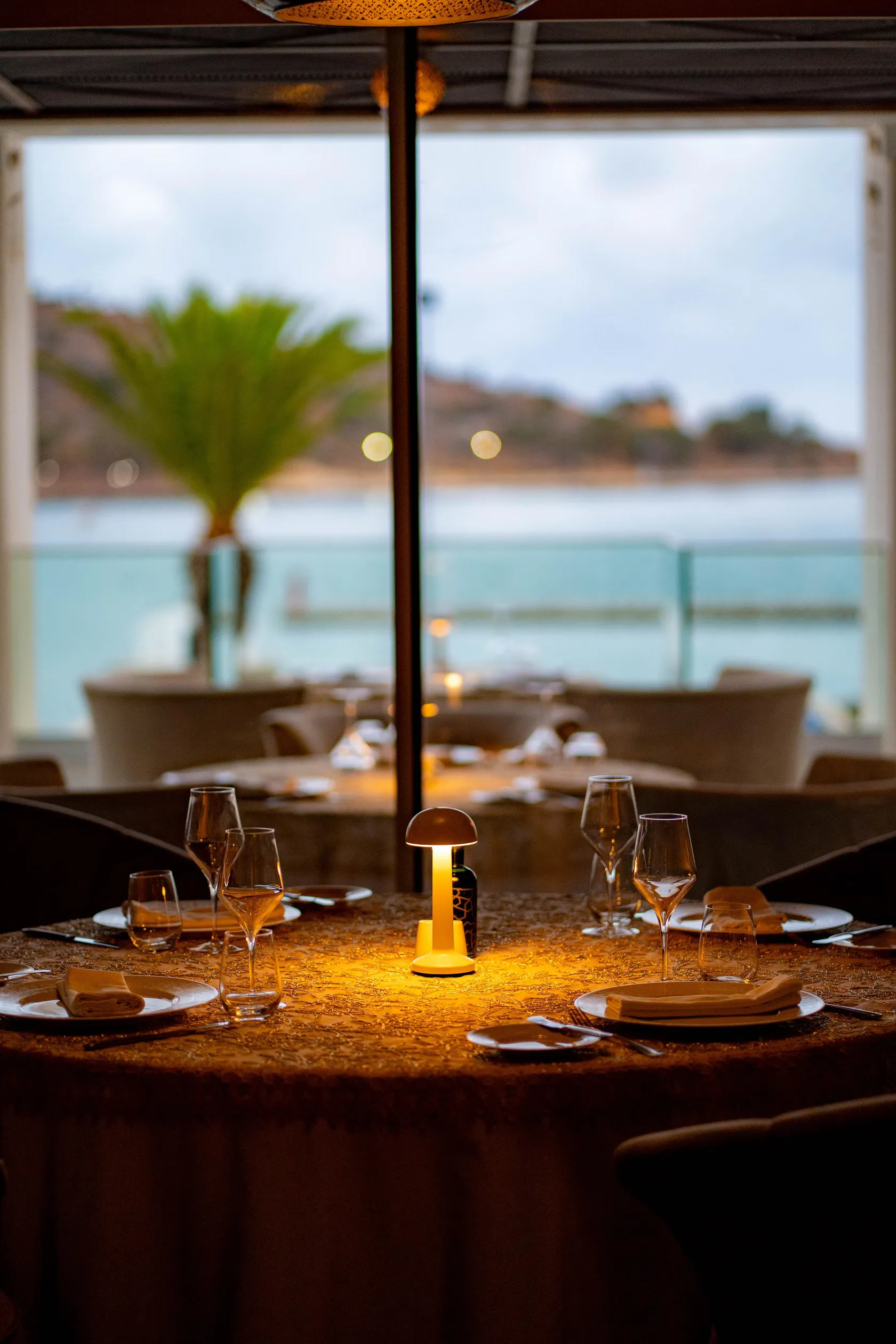 Table dressée avec verres à vin et bougie allumée, vue à travers une fenêtre donnant sur une terrasse et un paysage extérieur avec palmiers et mer