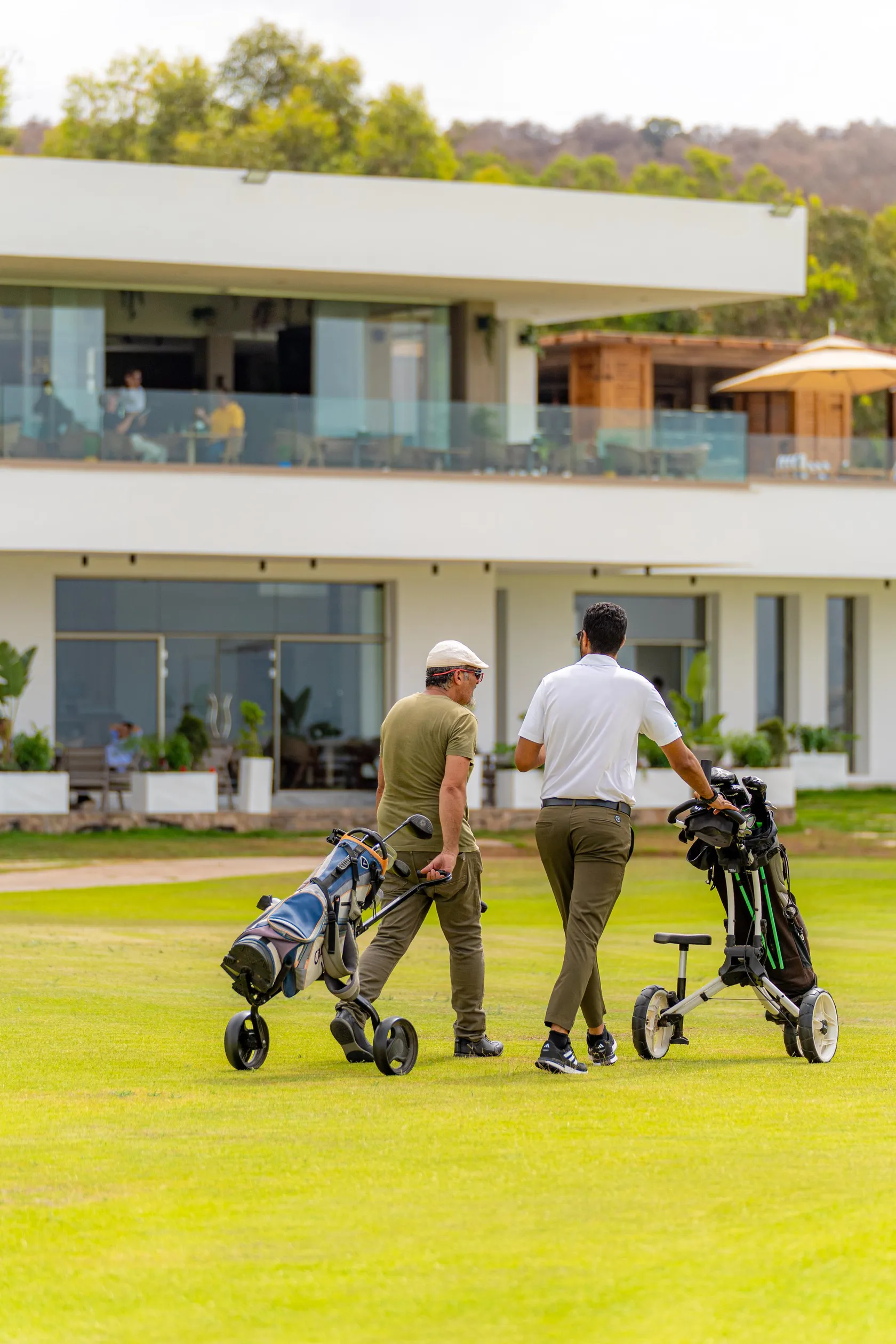 Two men walk on a golf course with club bags, in front of a modern building with large glass windows.