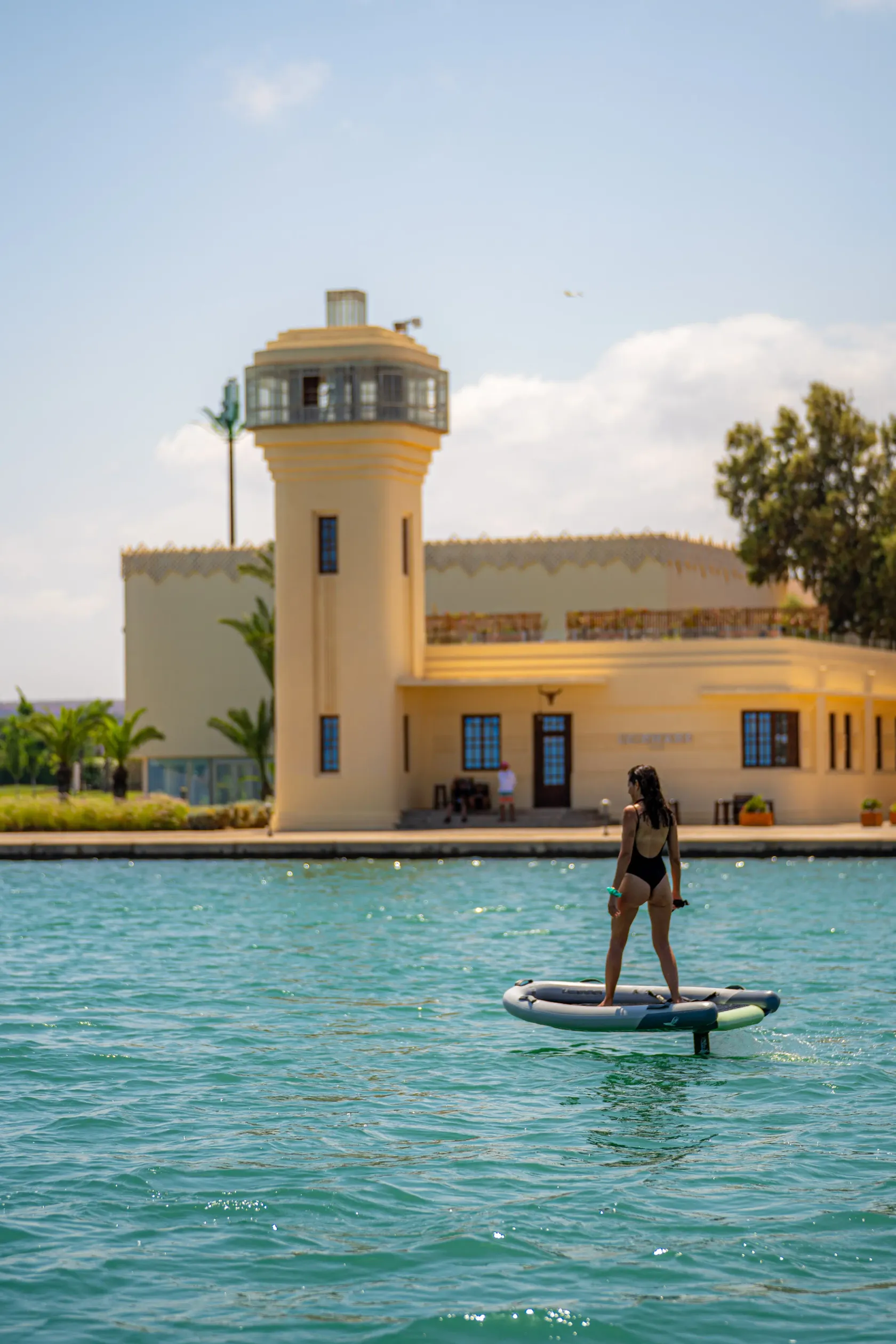 A person in a swimsuit is paddleboarding on a body of water in front of a yellow building with a tower.