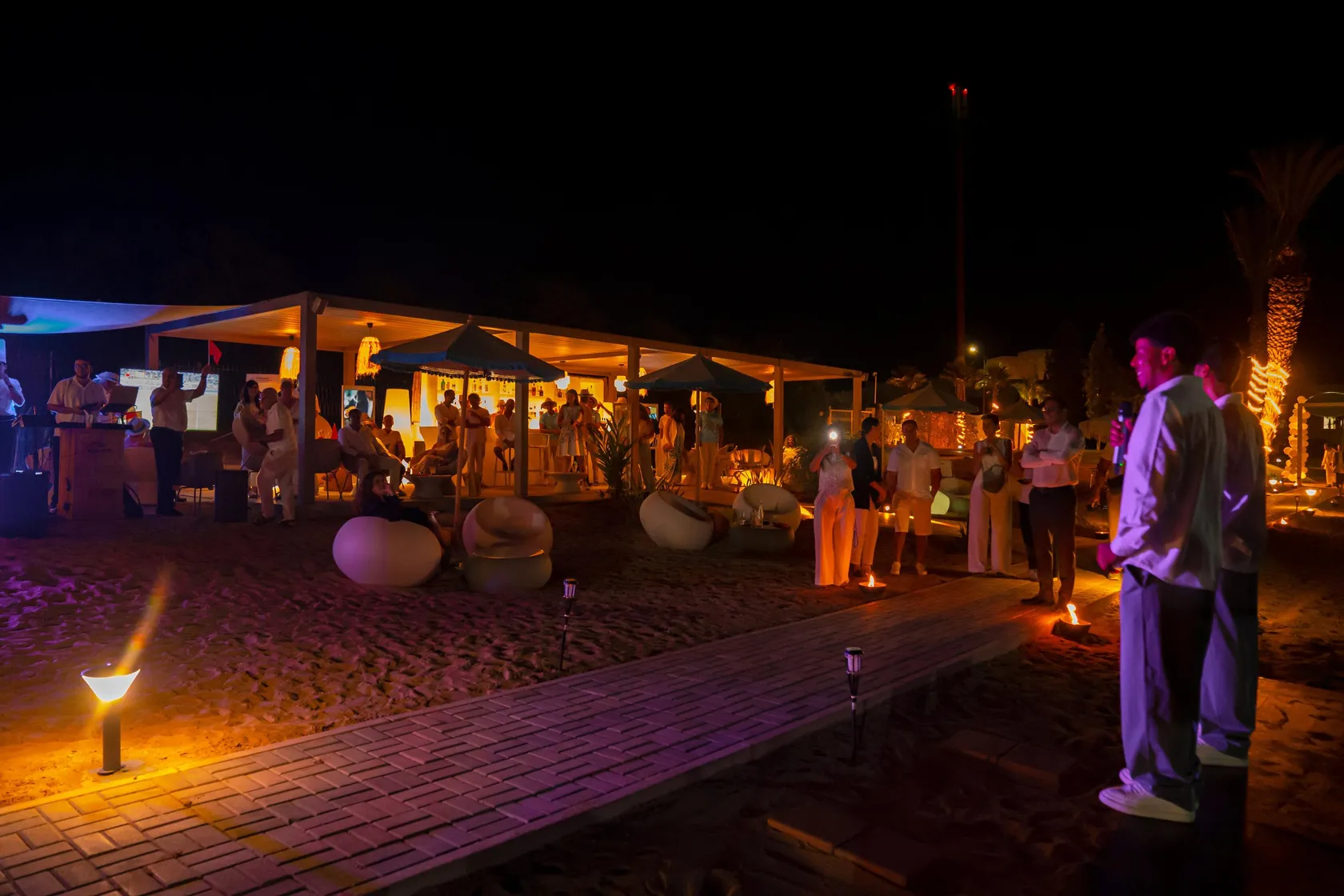 A group of people stands around a lit structure on a nighttime beach.