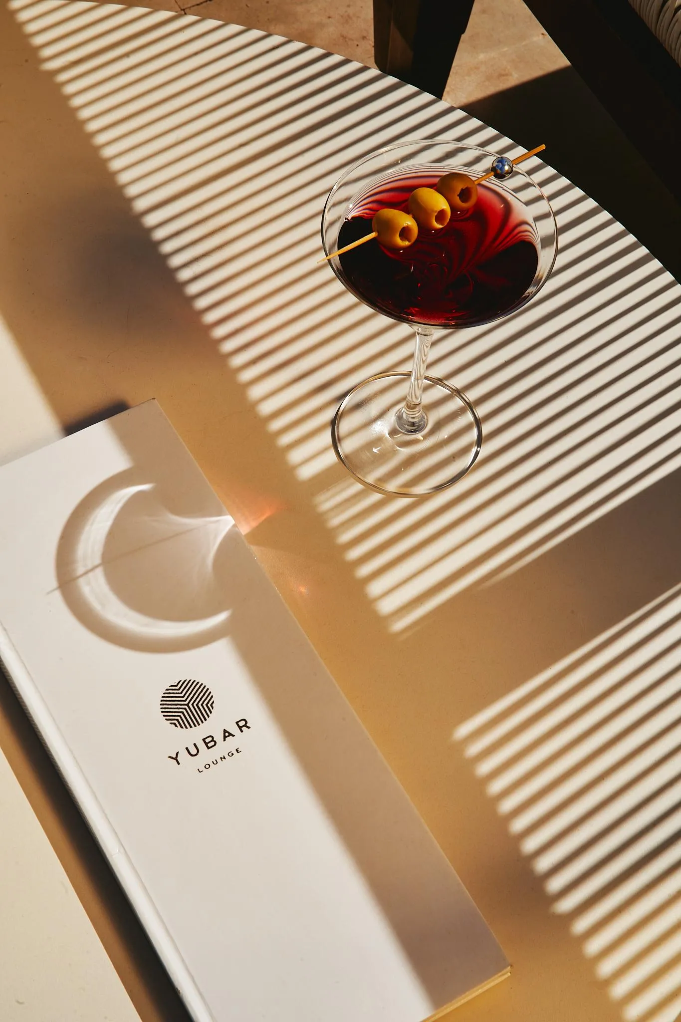 A glass of cocktail with olives and a straw, placed on a wooden table with a "YUBAR LOUNGE" menu next to it.