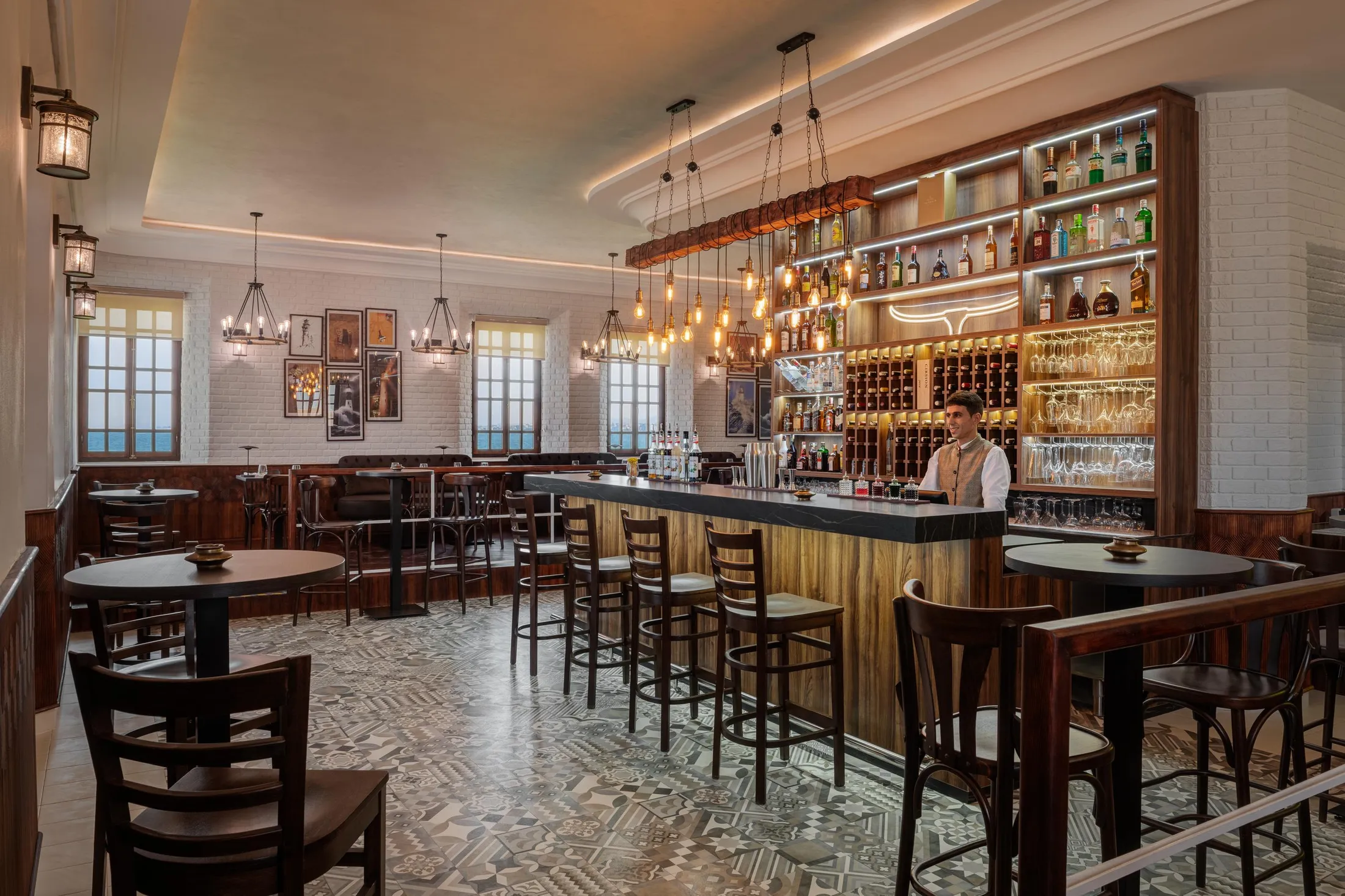 Un barman se tient derrière le comptoir d'un bar élégant avec des étagères remplies de bouteilles et des tables disposées autour