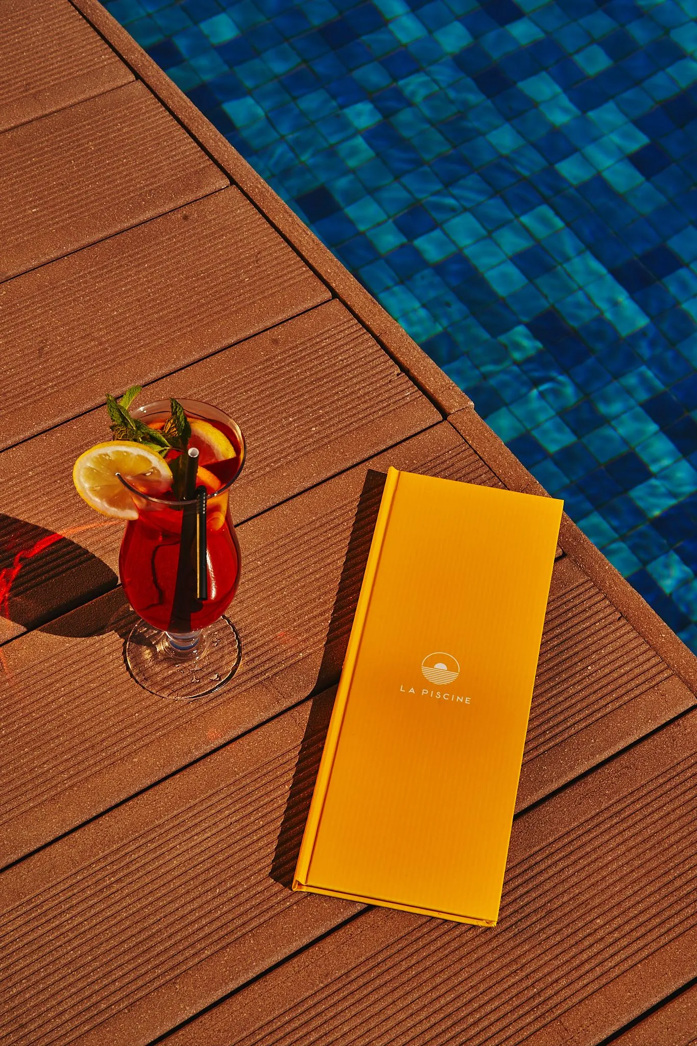 A glass of red drink with an orange slice and a straw, a yellow menu, and a blue pool in the background.