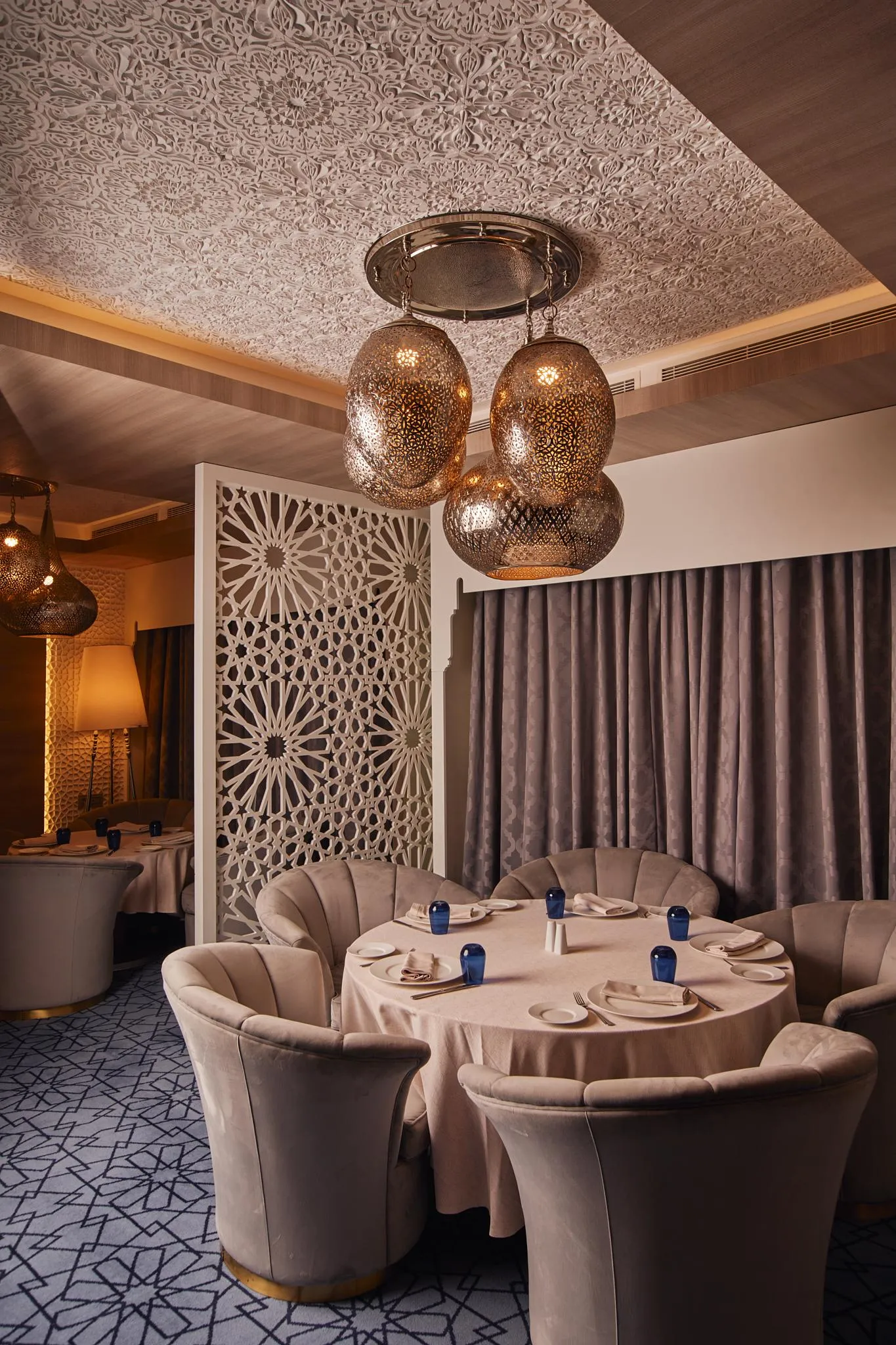 An elegant restaurant with round tables, padded velvet beige chairs, metal light fixtures, and a ceiling decorated with geometric patterns.