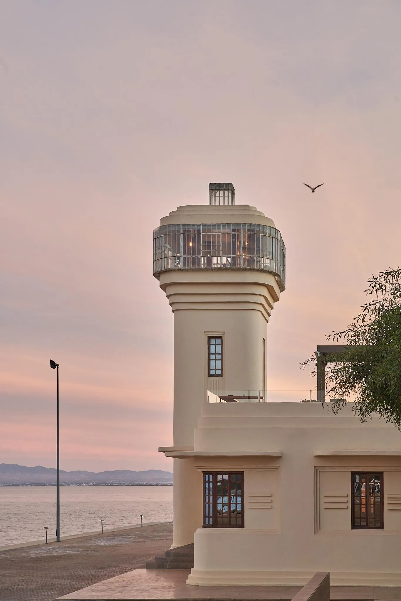 A modern lighthouse with a glass lantern at the top stands near a body of water at sunset.