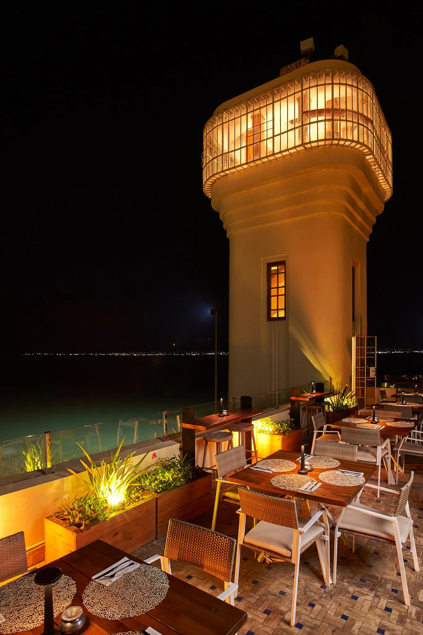 Restaurant terrace lit at night with tables, chairs, and plants, located near a lit cylindrical structure and a pool.