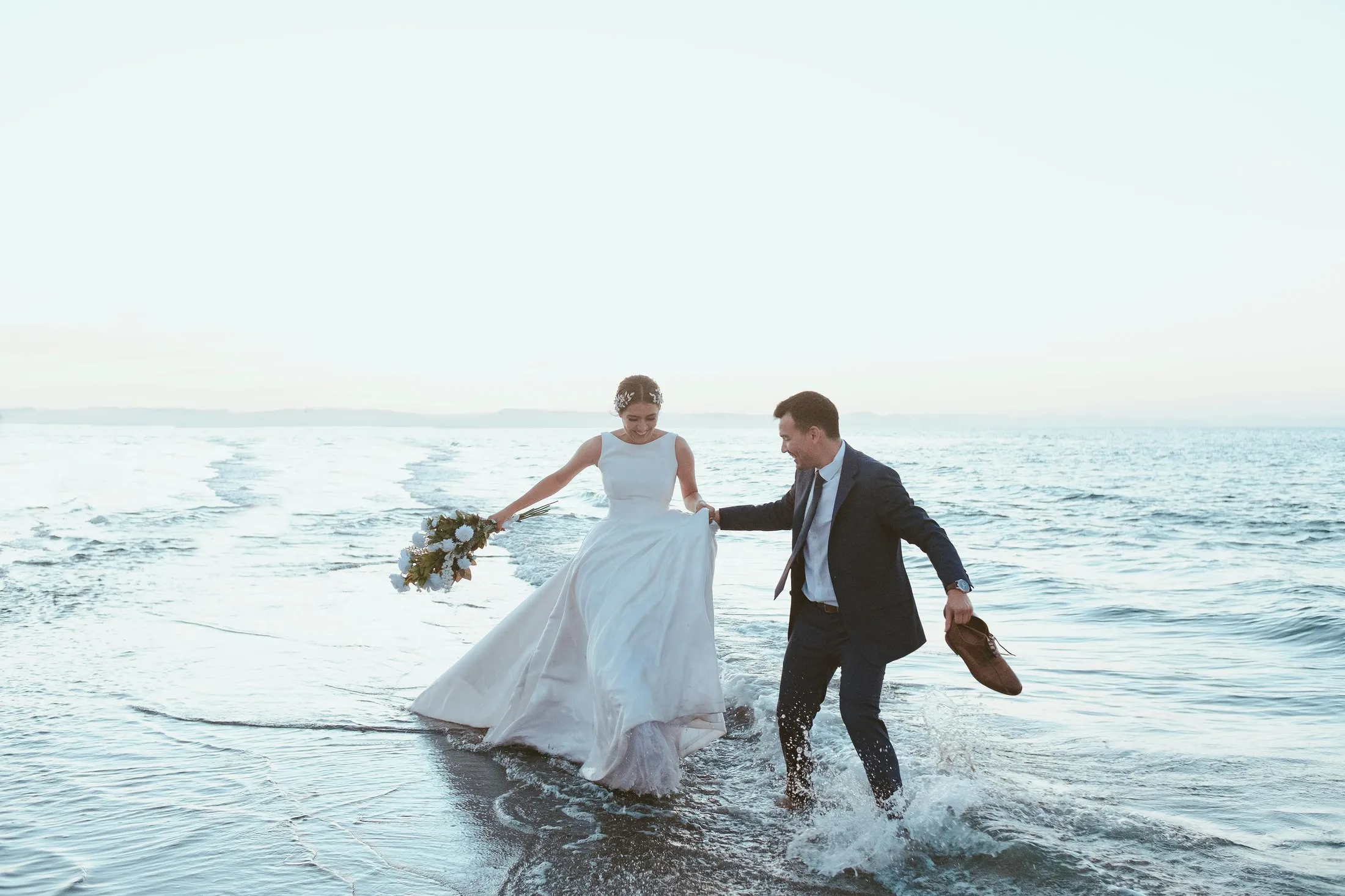 Una pareja camina descalza en el agua, la novia sosteniendo un ramo y el hombre sus zapatos.
