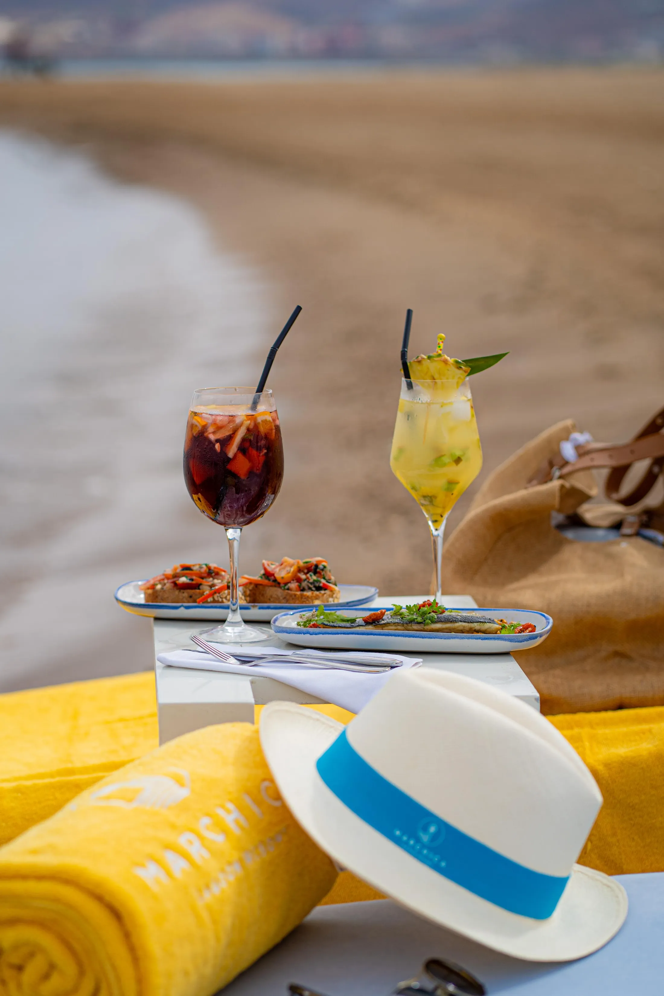 Two glasses of cold drinks with straws, plates of food, and a hat on a table near a beach.