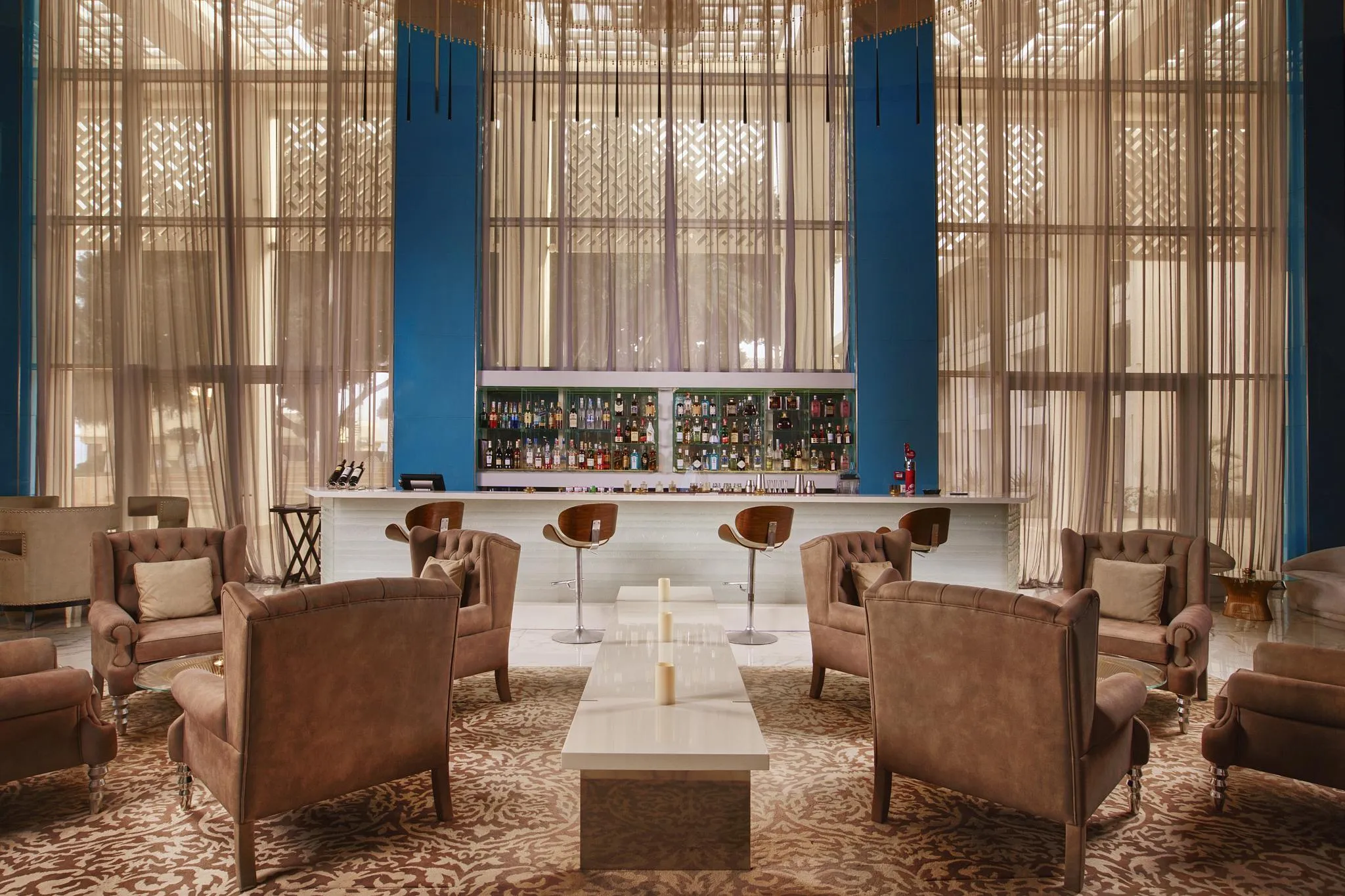 A living room with leather armchairs around a coffee table, facing a bar with shelves filled with bottles, behind large windows with sheer curtains.