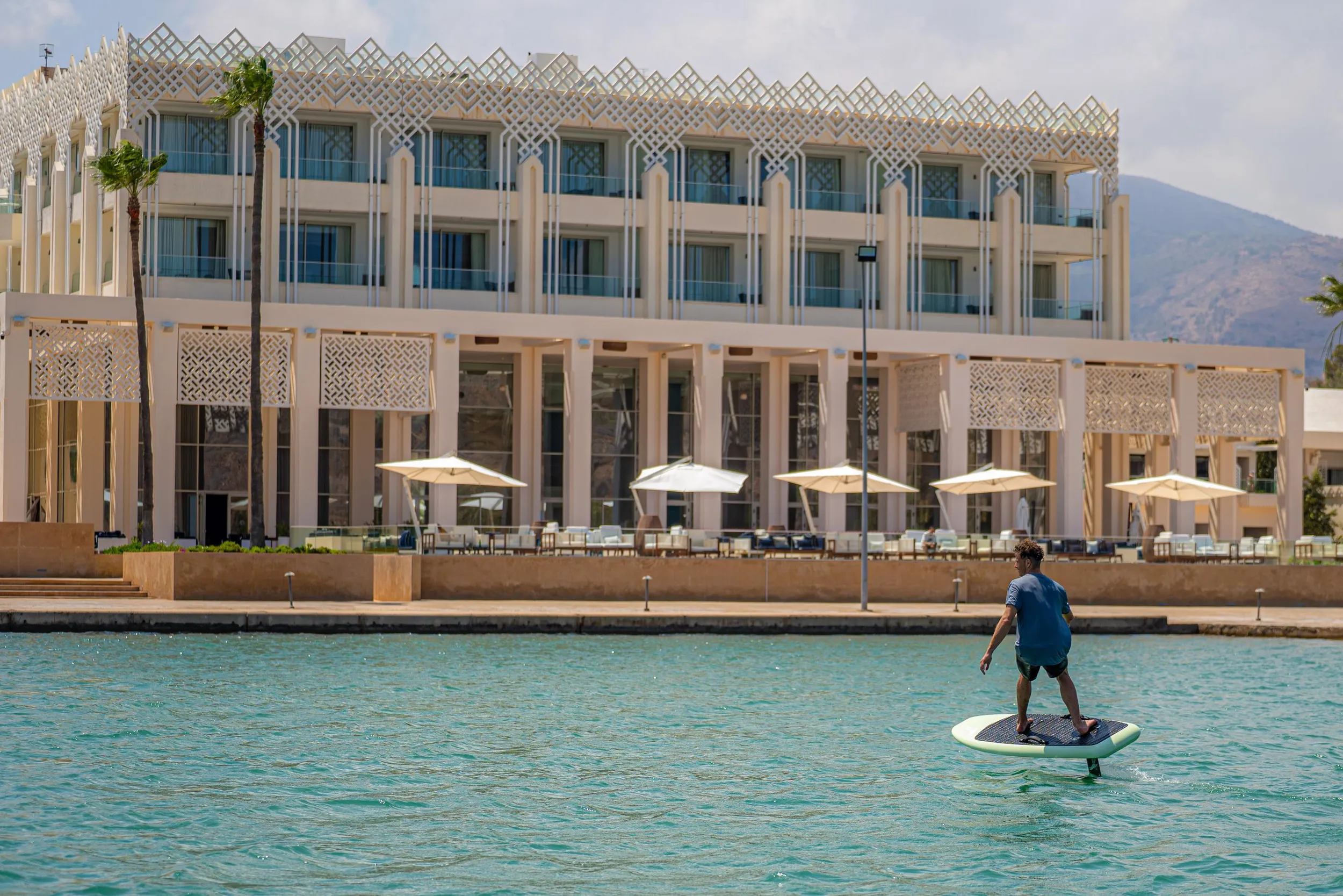 A man is paddleboarding on a body of water in front of a modern building with arches and balconies.