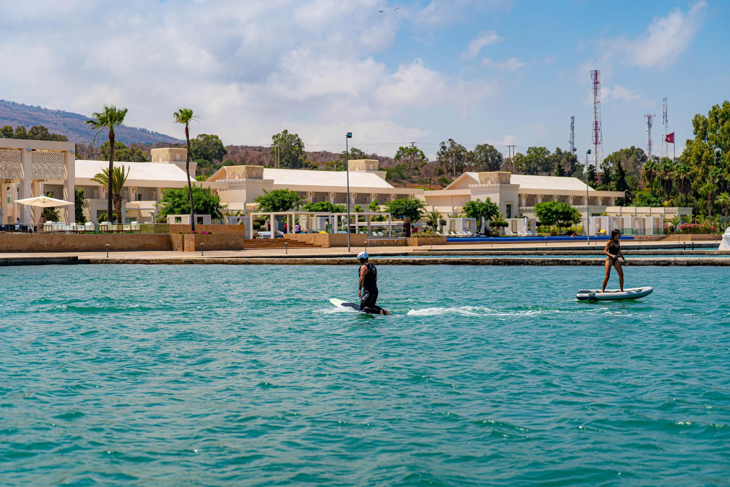 Two people are paddleboarding on a turquoise body of water in front of a white building with palm trees.