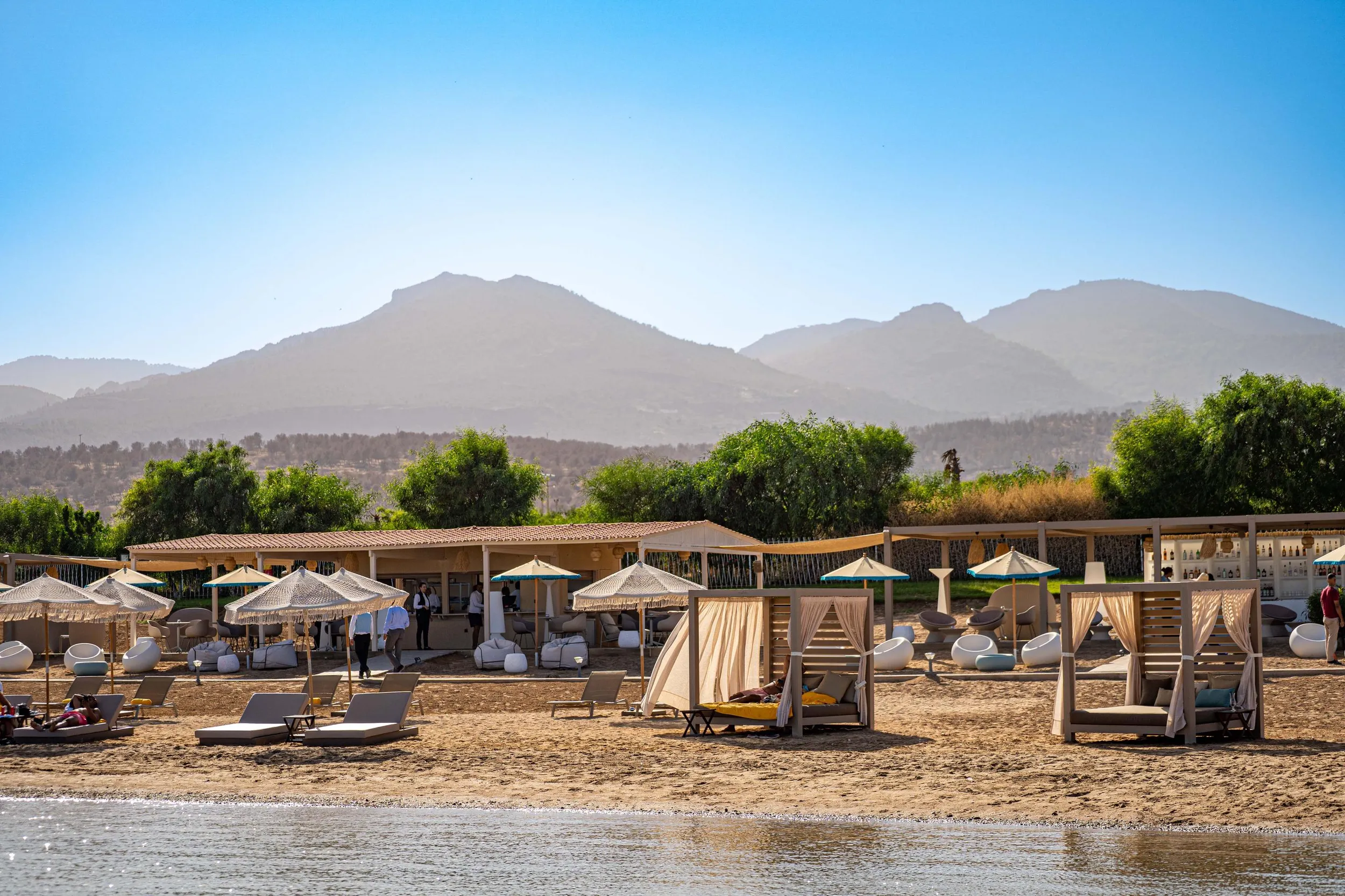 A beach with sun loungers, umbrellas, and wooden cabins, located at the foot of mountains with dense vegetation.