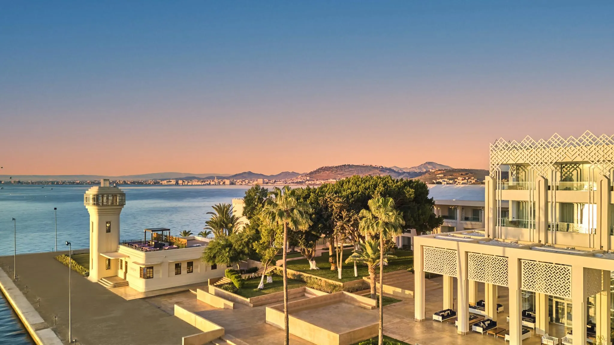 Vue d'un complexe architectural moderne en bord de mer avec des bâtiments blancs, une tour, des palmiers et une mer calme au coucher du soleil