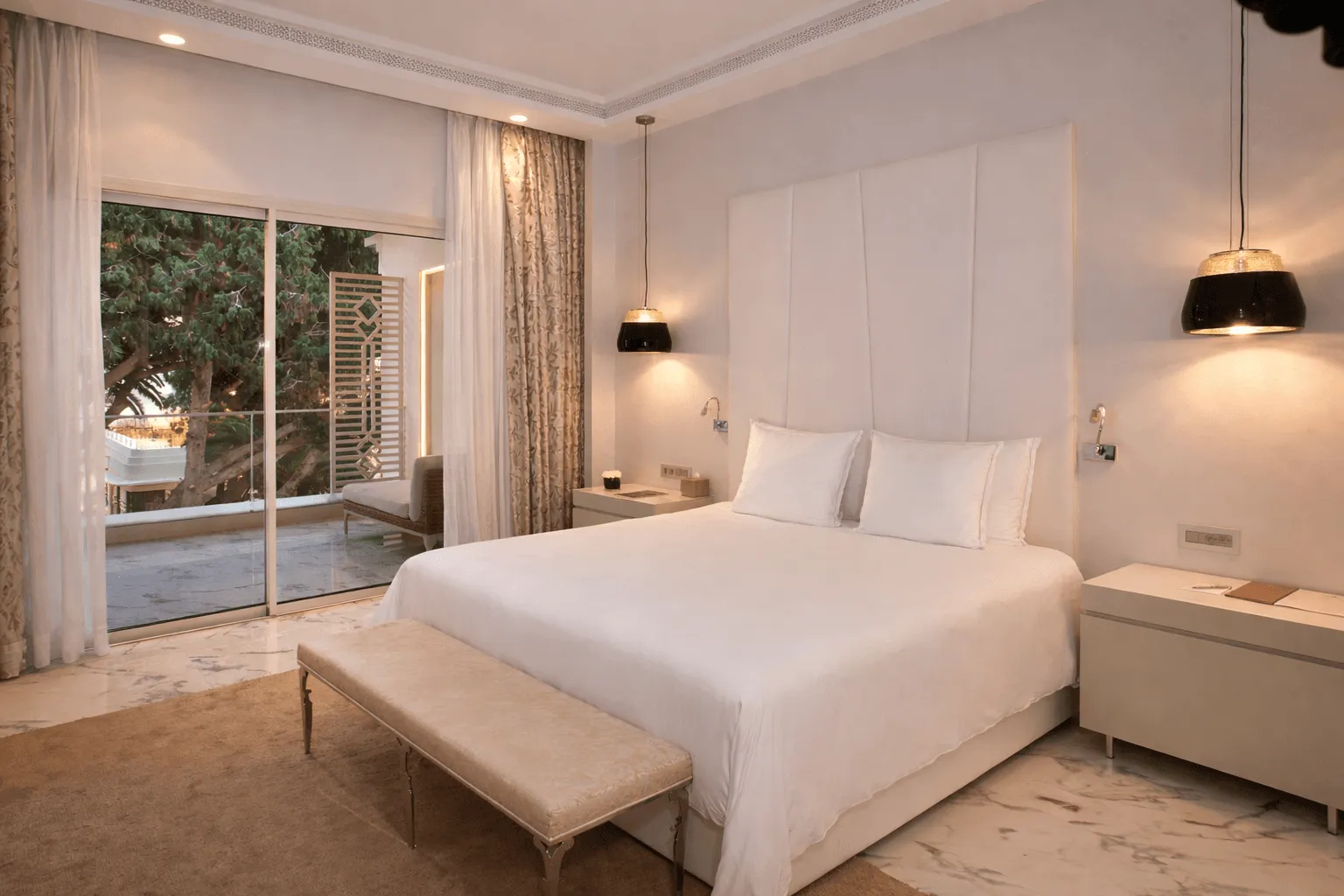 Habitación de hotel moderna con una cama doble, un banco al pie de la cama, lámparas colgantes y un gran ventanal que da a una terraza con vistas a árboles.