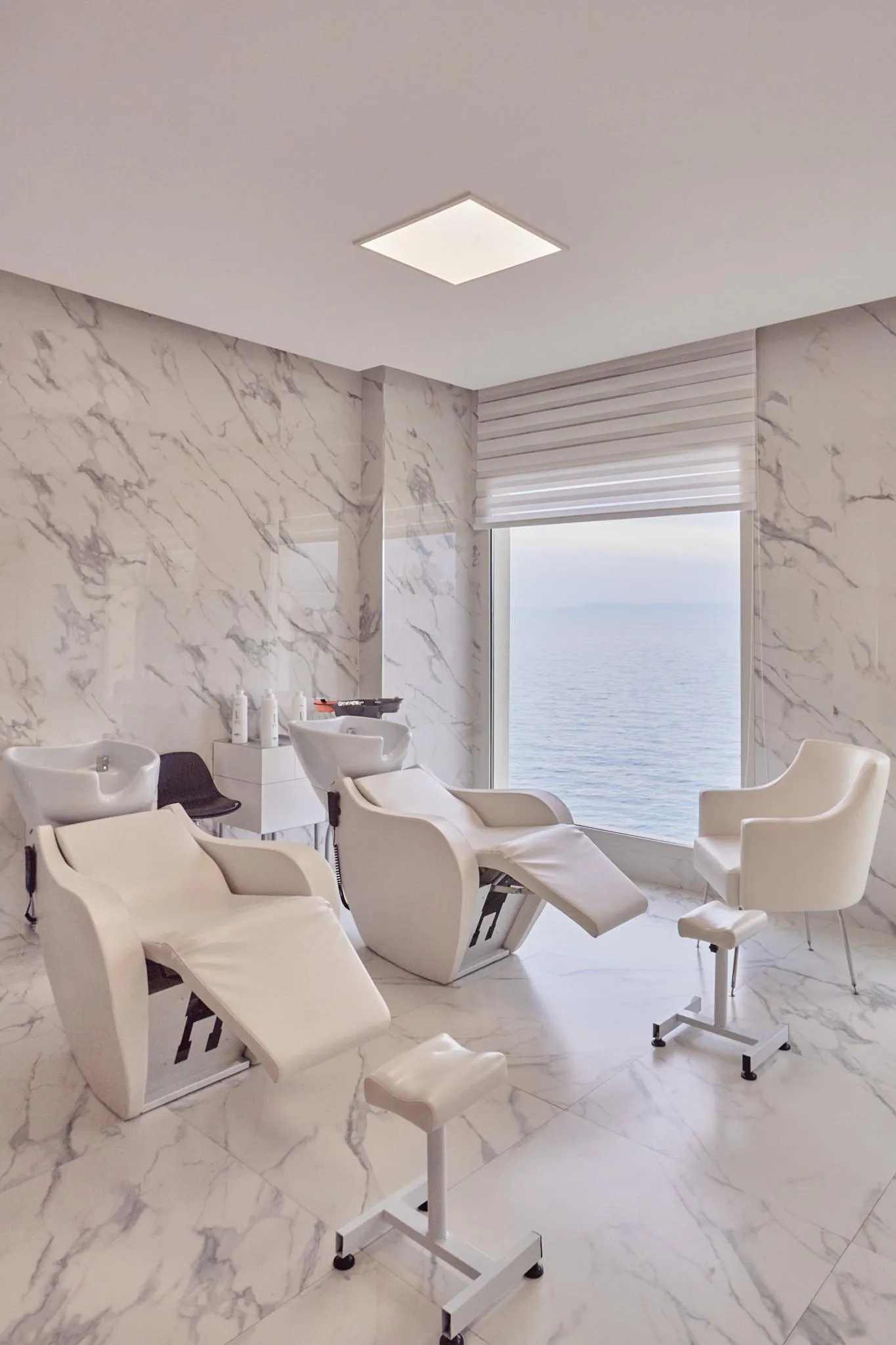 Hair salon with white hairdressing chairs and marble-covered white walls, offering a sea view through a large window.