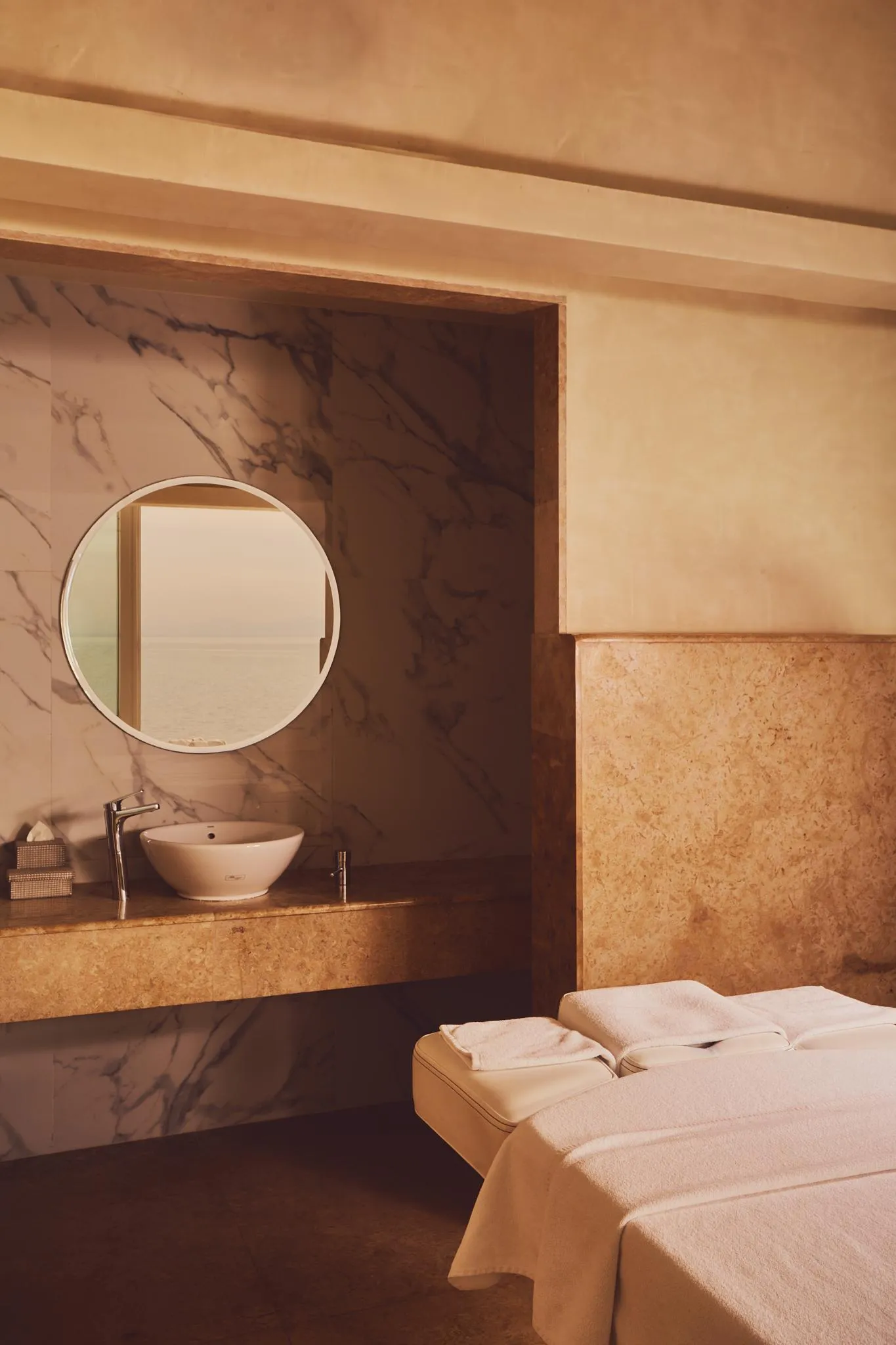 A sink built into a stone countertop with a round mirror above it, next to a massage table covered with white towels.