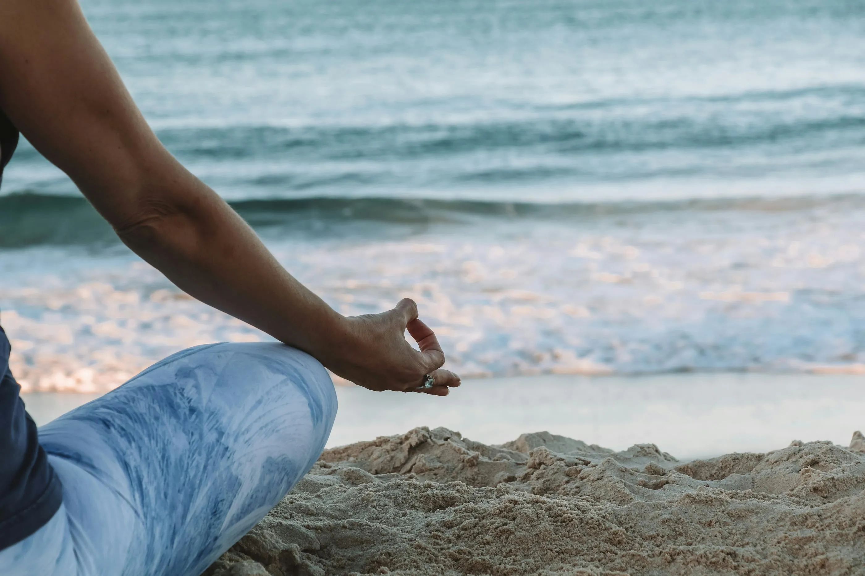Une personne en pantalon bleu clair est assise sur le sable près de la mer, la main posée sur le genou