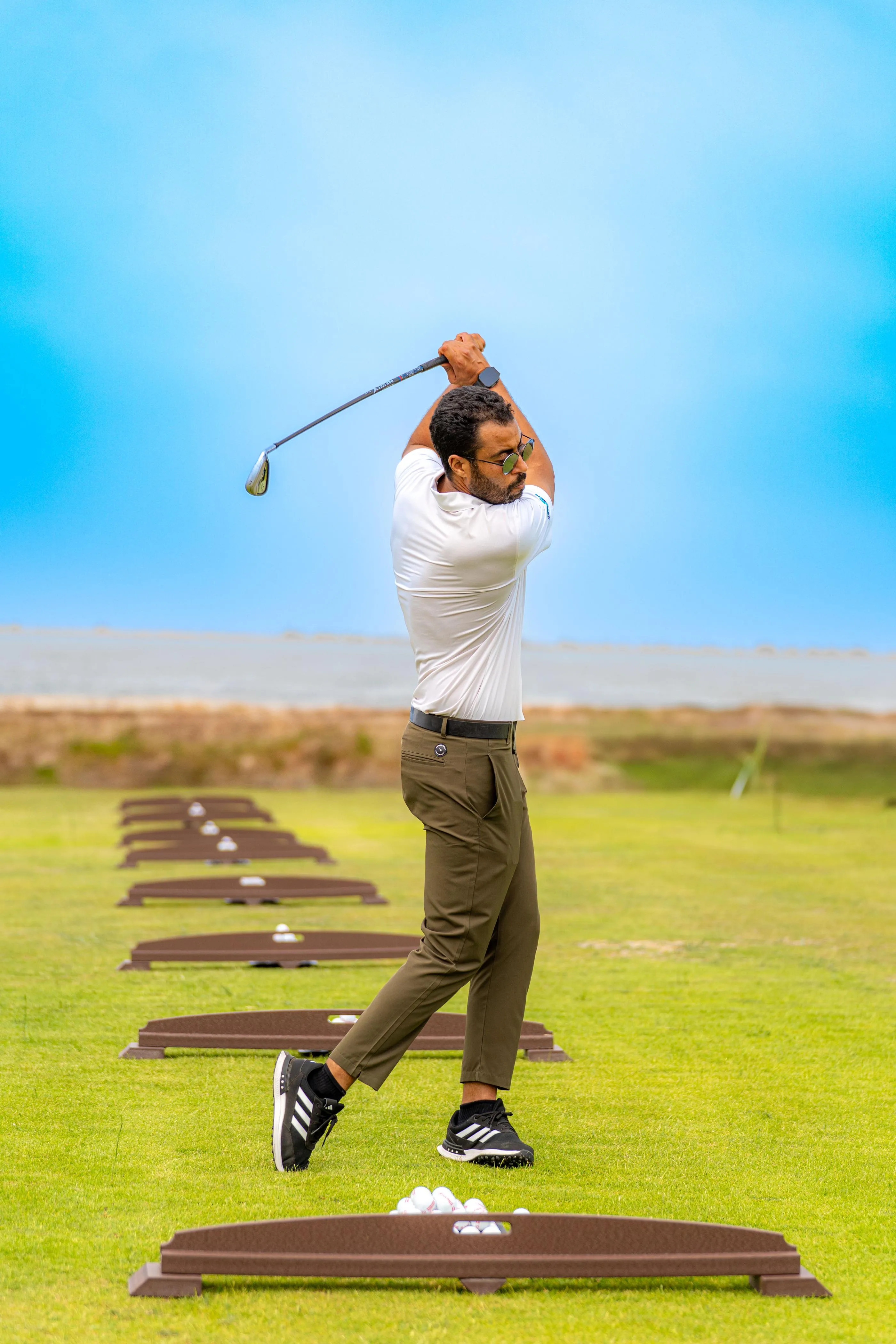 A man in a white polo shirt and green pants hits golf balls on an outdoor practice range.
