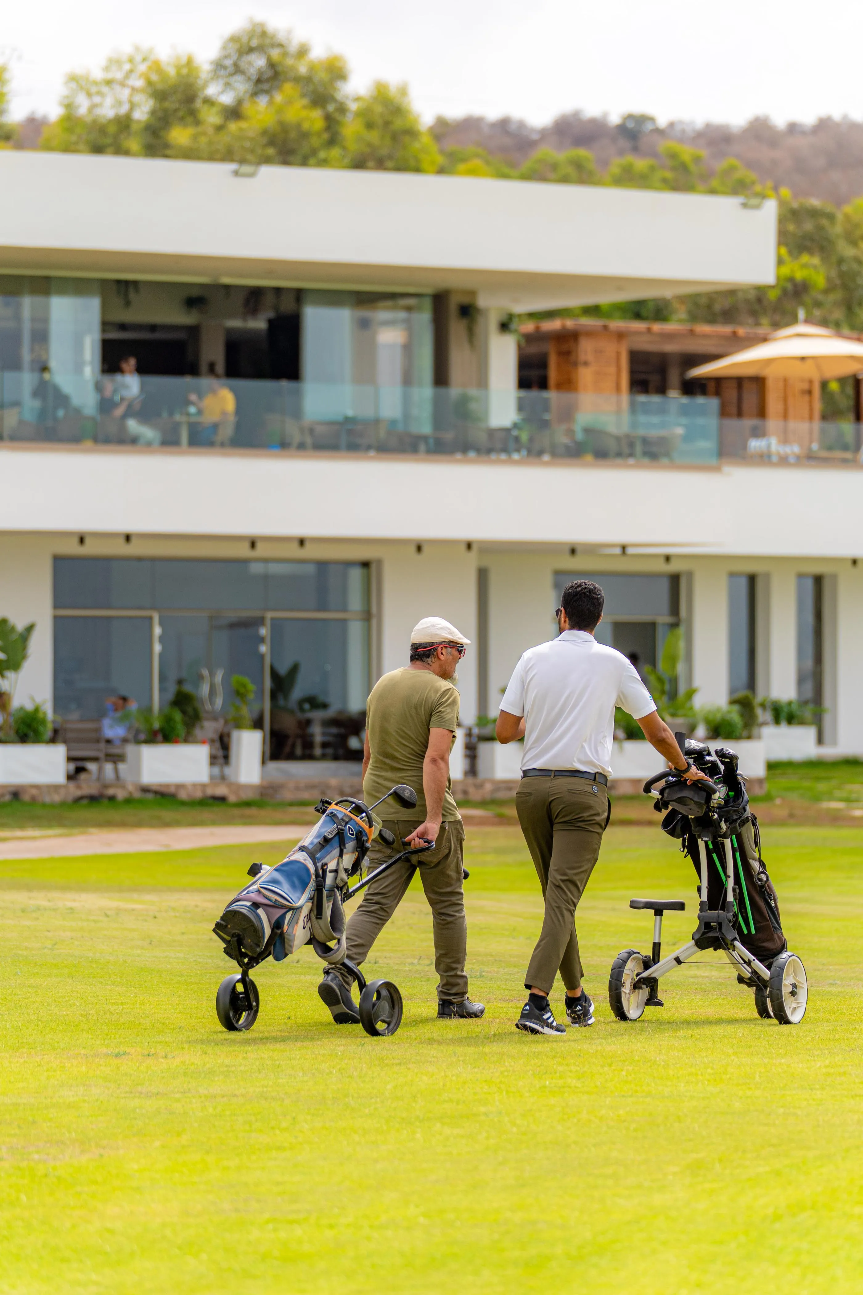 Two men walk on a golf course with club bags, in front of a modern building with large glass windows.