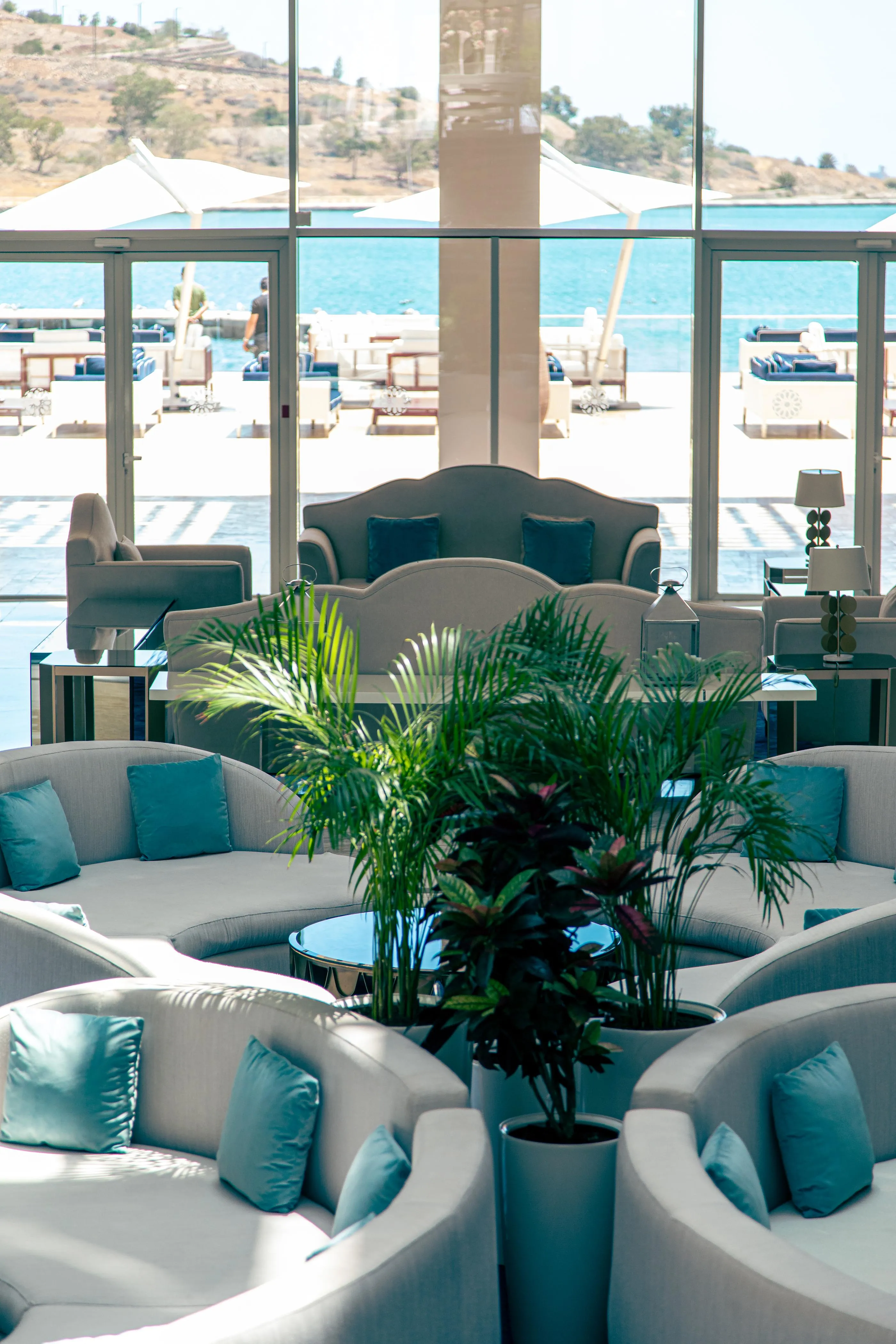 A living room with gray sofas and blue cushions, a green plant in the center, and a view of a pool and the sea through large glass windows.