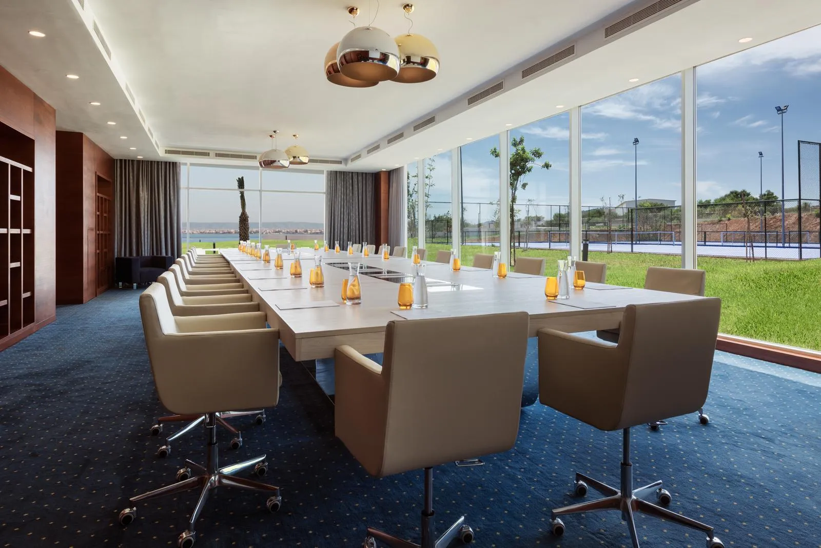Sala de reuniones con una larga mesa rodeada de sillas, vasos y candelabros sobre la mesa, grandes ventanas que ofrecen una vista a un terreno exterior