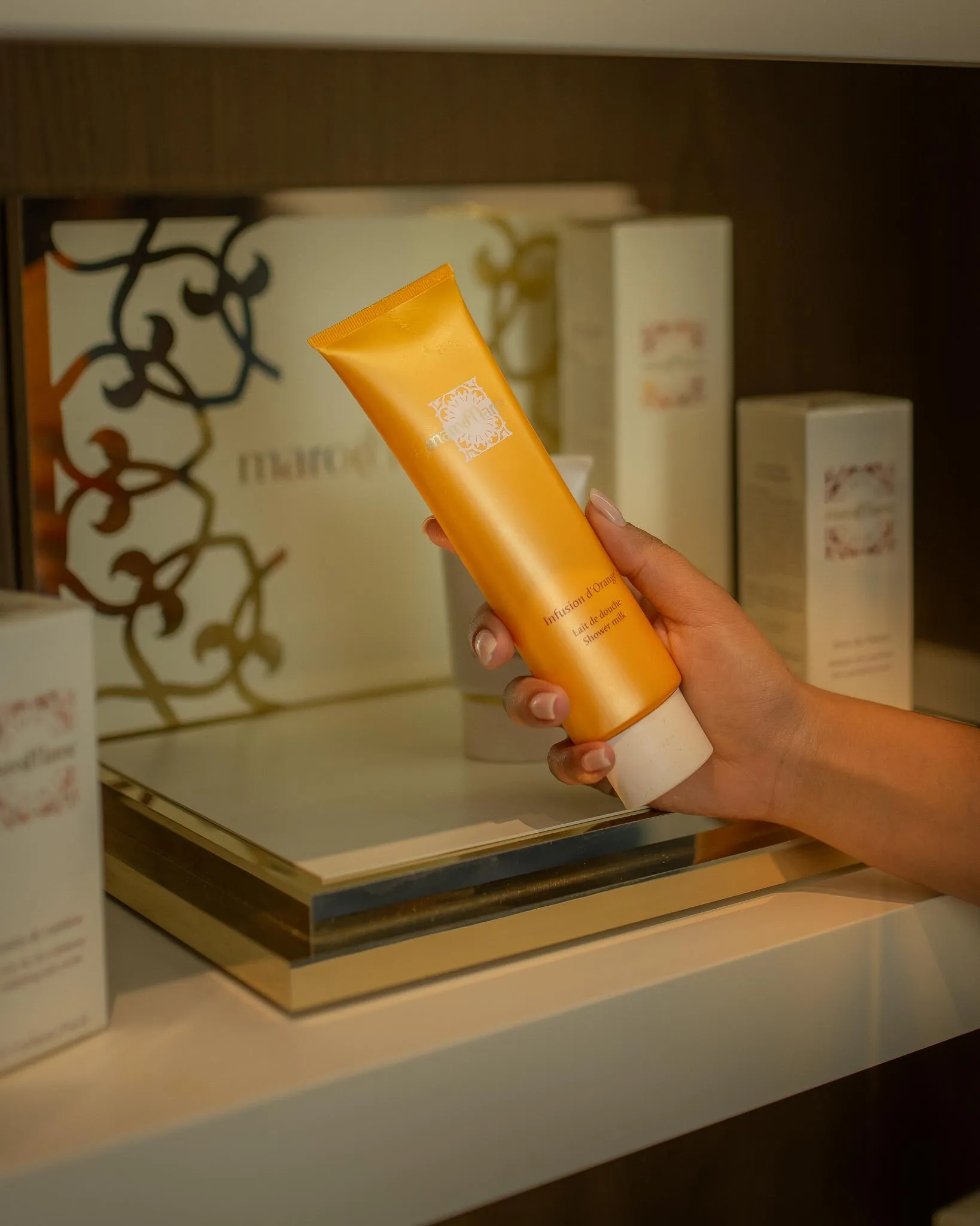 A hand holds a tube of cosmetic product in front of a shelf with other products.