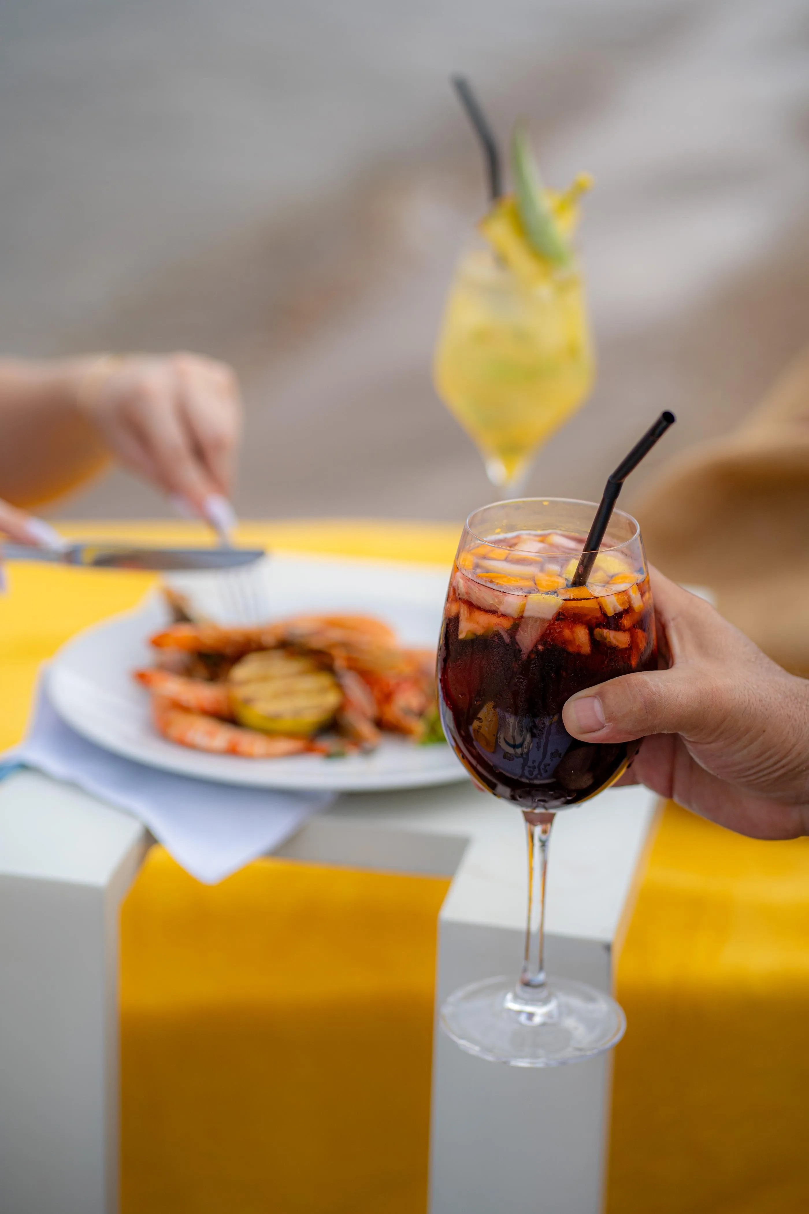 A person holds a glass of red drink with ice cubes and a straw, in front of a plate of food and a yellow table.