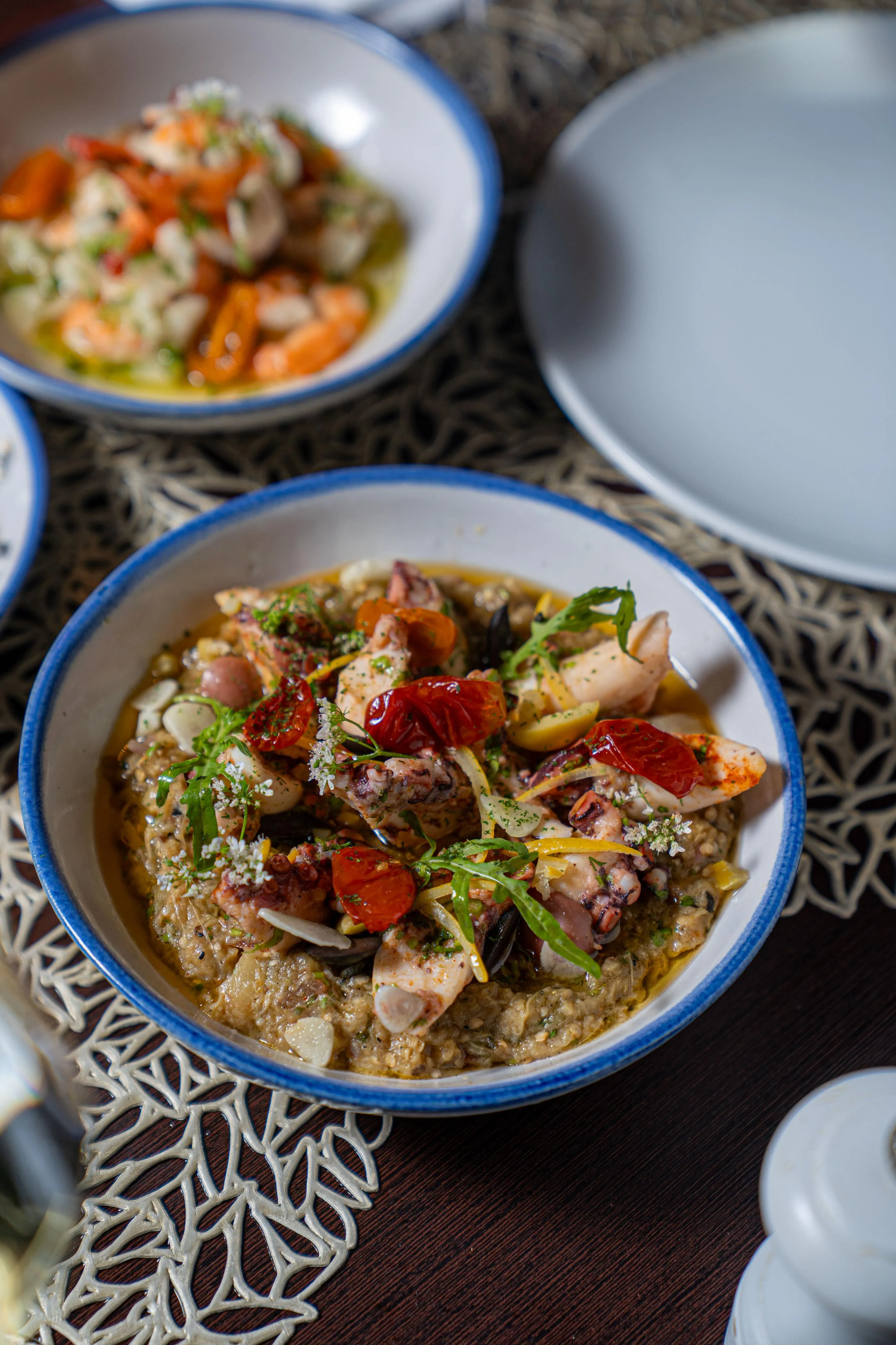 Bowl of food containing chicken, vegetables, and grains, garnished with herbs and cheese, placed on a patterned tablecloth with other bowls in the background