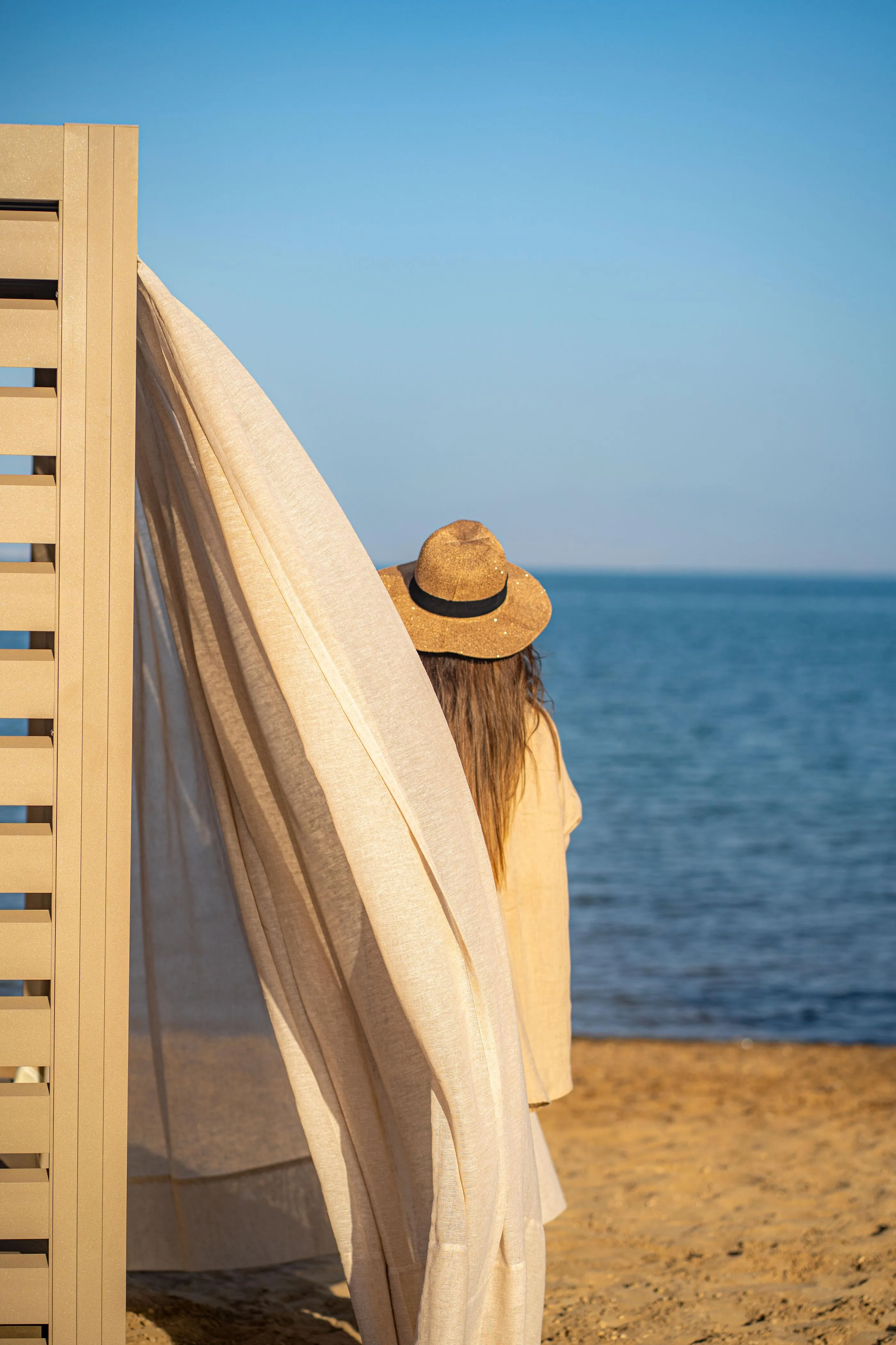 A person wearing a hat looks at the sea from a beach, a white sheet hanging from a wooden structure next to them.