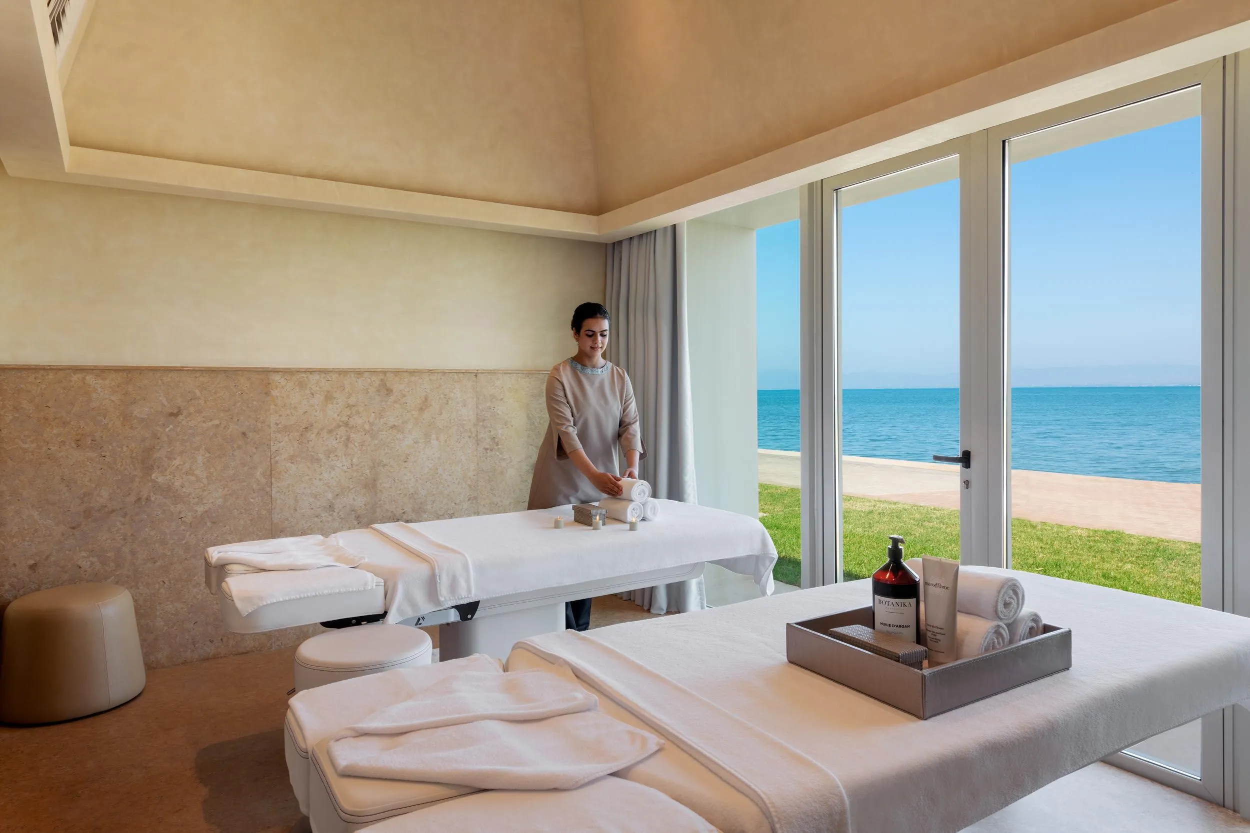 A person in work attire prepares a massage table in a room with a sea view.