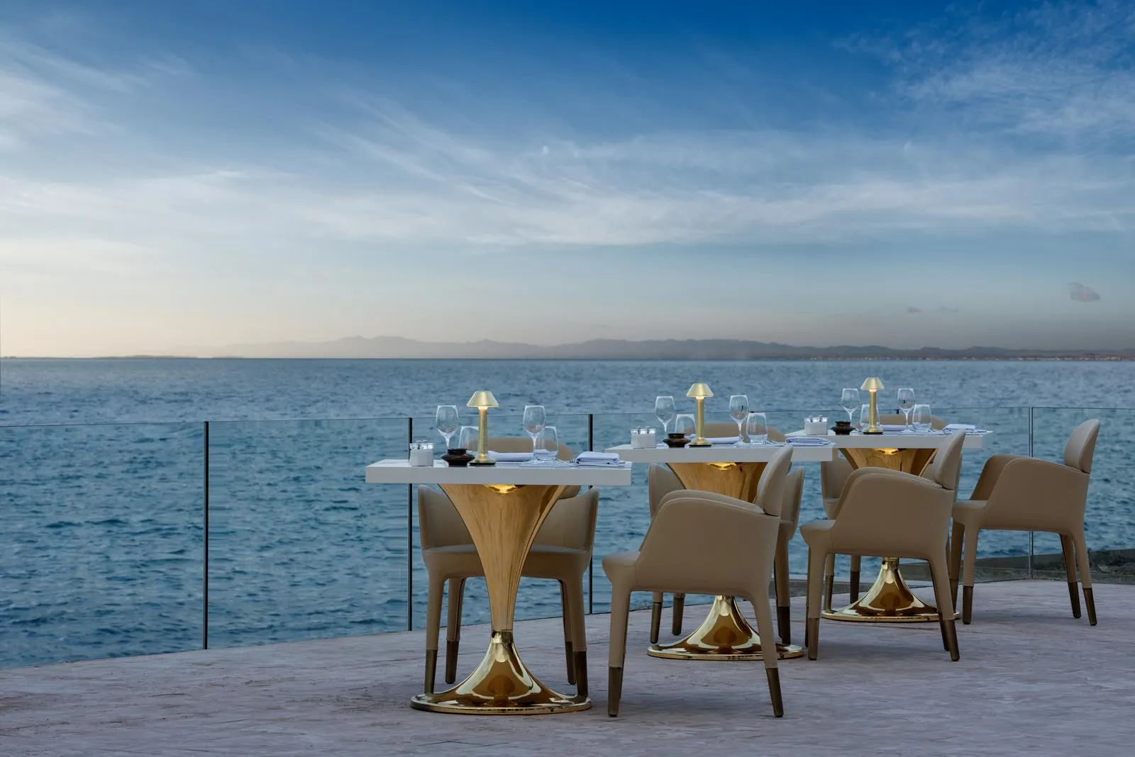 Mesas y sillas dispuestas en una terraza frente al mar con vasos y servilletas, bajo un cielo despejado