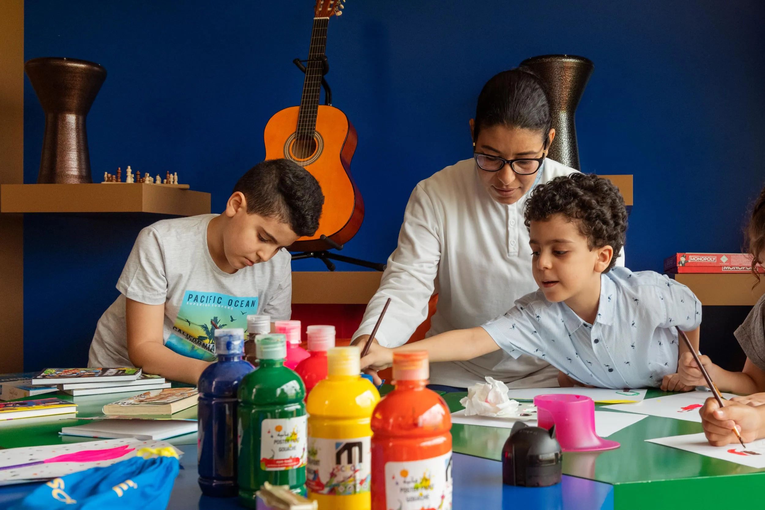 Three people are sitting around a table with paint bottles and brushes, drawing on sheets of paper.